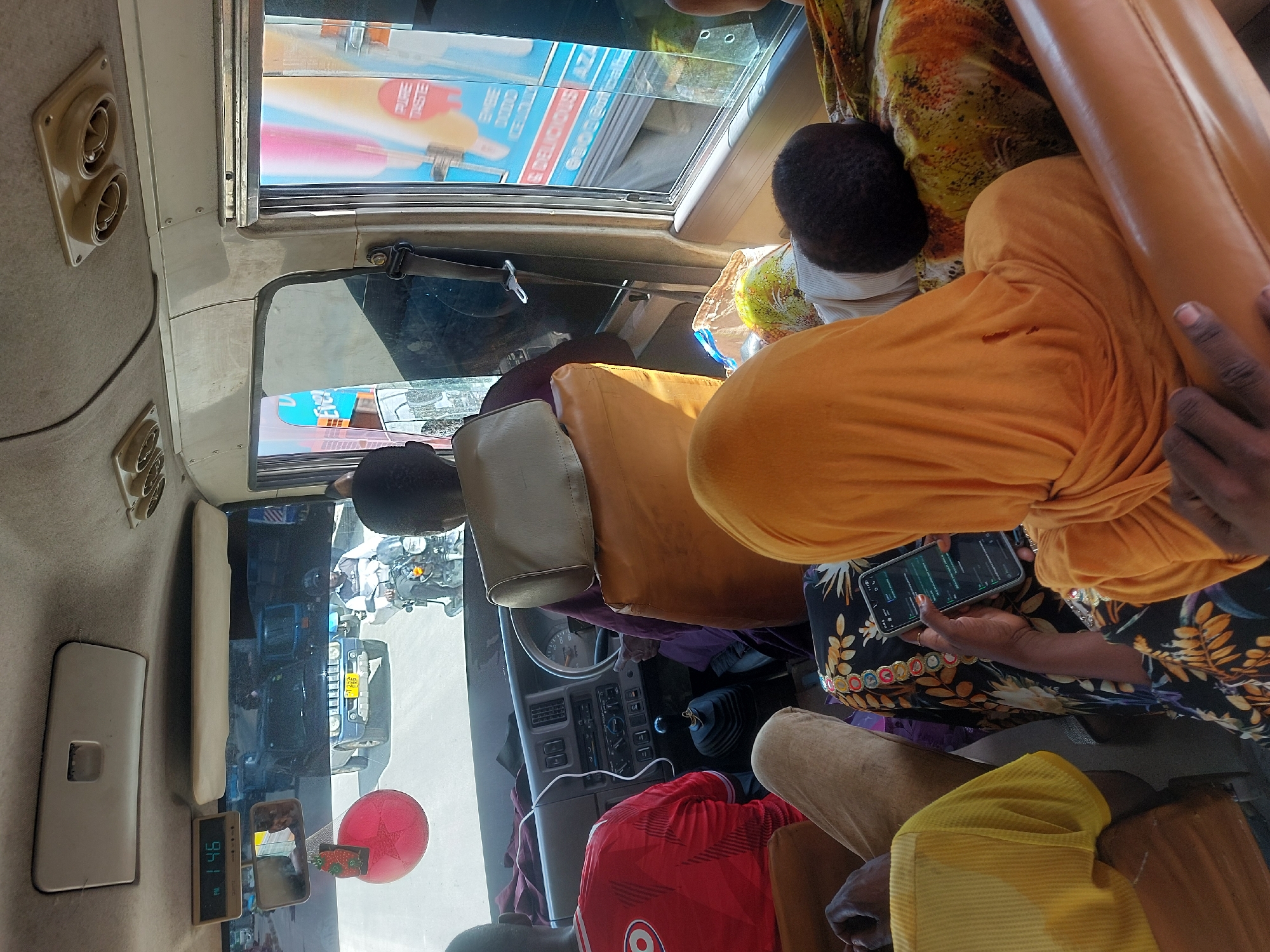 The image captures the interior of a busy public transport vehicle, likely a "daladala" in Dar es Salaam, Tanzania, during the early afternoon. The perspective is from inside the vehicle, looking towards the front and out the windows.
Several passengers are visible. In the foreground, a person wearing a vibrant orange garment and a black and yellow patterned dress or shirt is seated, holding and looking at a smartphone. Their dark-skinned hand and nails are visible as they operate the device. Directly behind them, the head of another passenger, wearing a dark cap and a light-colored shirt, is seen. Further towards the front of the vehicle, partially obscured, is a person in a red t-shirt with a white circular logo, possibly the driver. Another person's legs, clad in light brown trousers and a yellow garment, are visible in the bottom right corner.
The interior of the vehicle features beige/off-white upholstery and trim. Overhead, there are two sets of air vents and a rectangular light fixture. A small digital clock displays "PM 1:46". A red spherical decoration or air freshener hangs near the front windshield, and a seatbelt is buckled to the side. The dashboard with its controls and steering wheel is partially visible, along with a gear stick. A clear plastic bag containing what appears to be green fruit rests on a seat.
Through the windows, the bright, sunny weather is apparent. The outside world reveals other vehicles, including a blue SUV or pickup truck with a partially visible license plate starting with "T...", and several motorcycles. Buildings and various advertisements are also visible. Text on an advertisement through the upper window includes "PURE TASTE," "DELICIOUS," "AZ," "ORGE," and "CRUM." The scene suggests an active urban environment. No strong emotions are discernible on the passengers' faces; they appear to be engaged in their travel or personal activities.