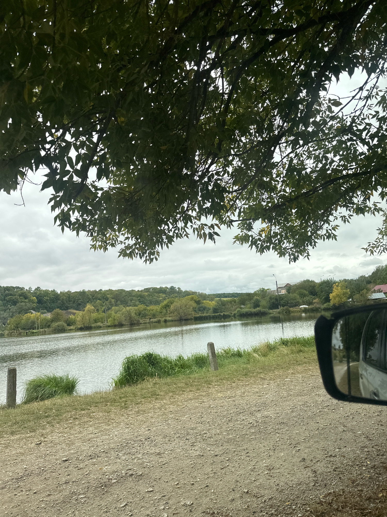 The image depicts a natural scene near the city of Makiv, Ukraine, on an overcast day. In the foreground, a dirt road is visible, with patches of dry grass and a small wooden post. To the left, a body of water, likely a lake or river, stretches across the middle ground, with its surface rippled. Lush green vegetation lines the opposite bank, consisting of trees and shrubs. In the distance, rolling hills covered in a dense forest form the horizon. The sky is filled with heavy, gray clouds. Partially visible in the lower right corner of the image is a car's side mirror, reflecting a portion of the car. Overhead, the branches and leaves of a large tree with dark green foliage obscure a significant portion of the upper frame.