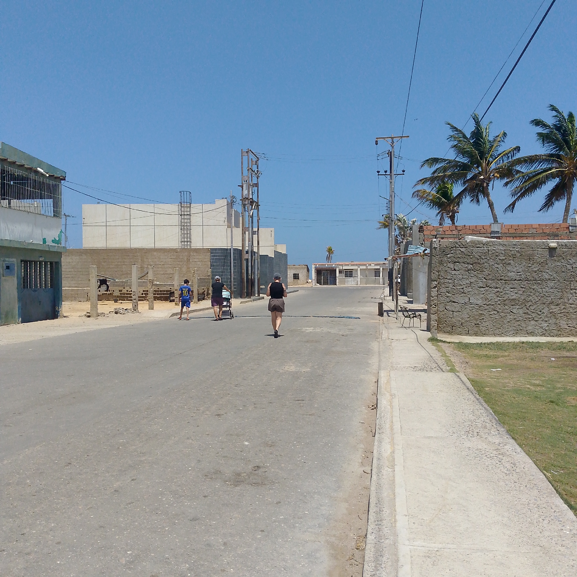 A street in Villa Marina, Venezuela. A boy and a woman with a stroller walk down the left side of the street, and another woman walks down the middle of the street away from the camera. There are unfinished buildings on the left and right side of the street, with electrical poles overhead. Three palm trees are visible on the right side.