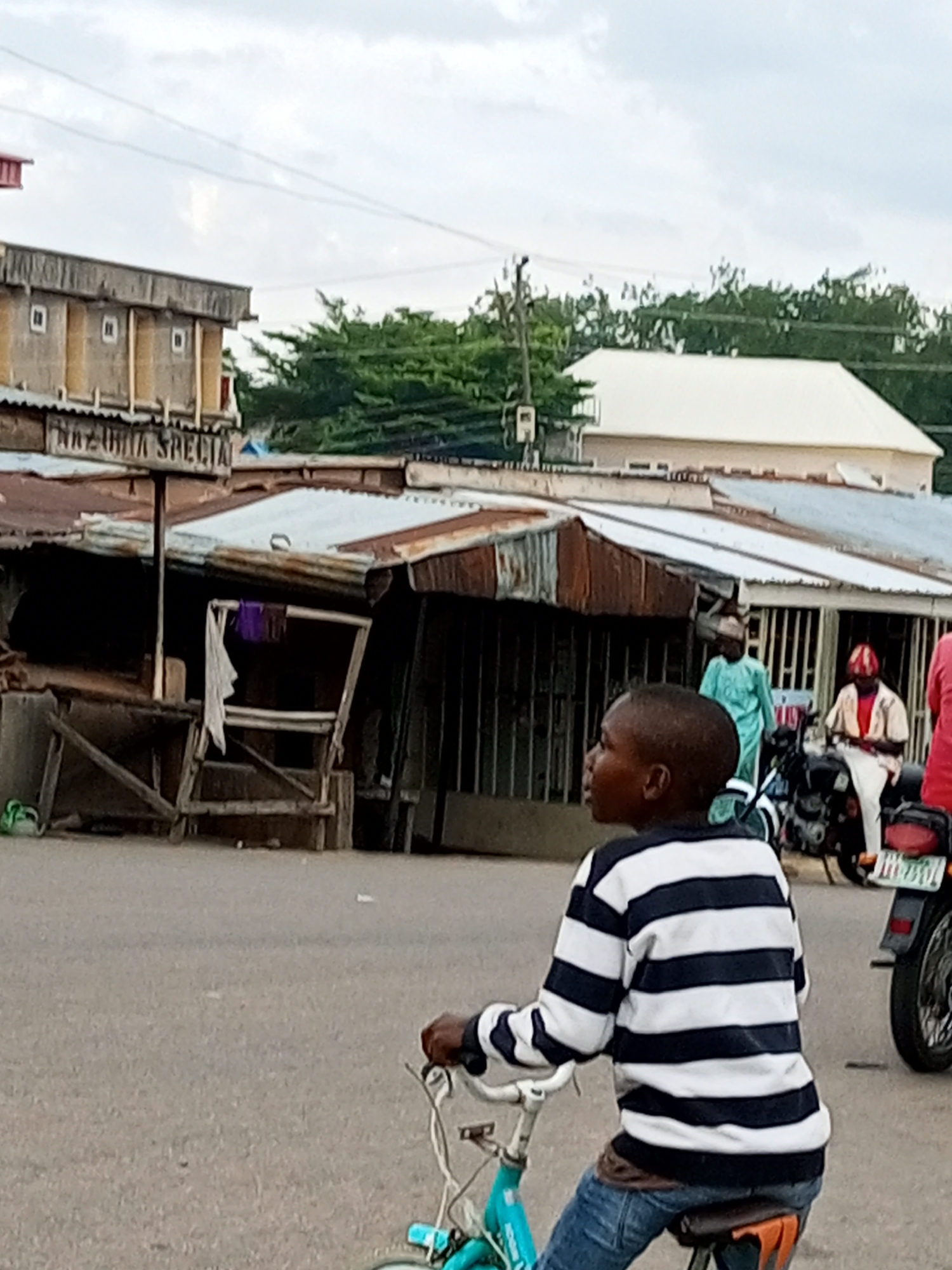 An outdoor, medium shot captures a young boy in the foreground riding a turquoise bicycle. He is wearing a black and white striped long-sleeved shirt and blue jeans. His head is turned to his left, and he appears to be looking ahead. The background shows a street scene with several buildings and people. To the left, a two-story building with a light-colored facade and visible windows stands. Below it are corrugated metal structures with wooden supports and a sign that reads "NAZURITA SPECIAL". In the middle ground, a person in a light blue garment walks away from the camera. To the right, another person sits on a motorcycle, wearing a red head covering and a light-colored shirt. The road surface is grey asphalt. The sky is overcast with a cloudy sky.