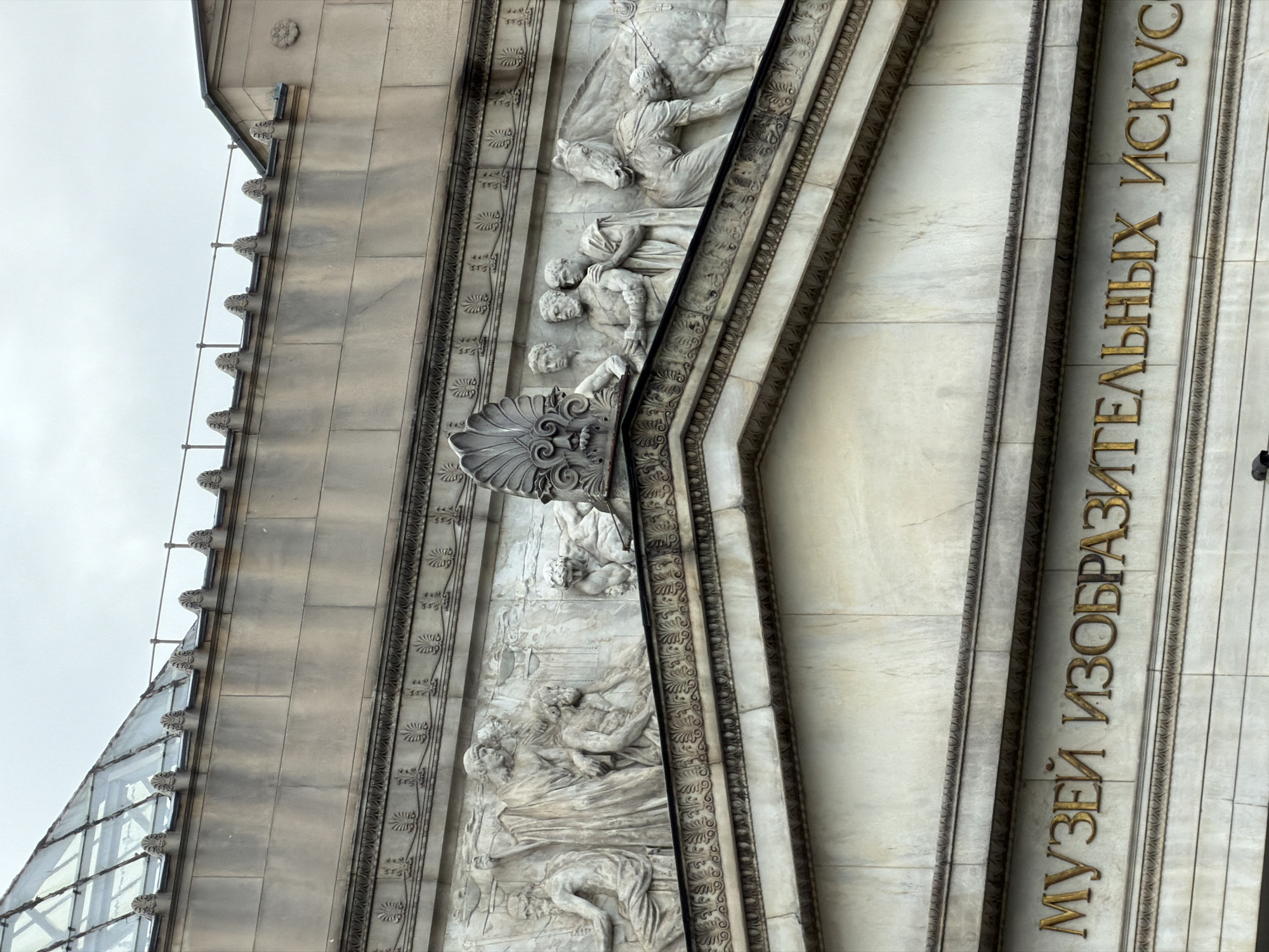 This media file captures an upward-looking view of a richly detailed building facade, identified as a museum in Moscow, Russia. The structure is built with light-colored stone, possibly marble, exhibiting a weathered yet majestic appearance under an overcast sky, suggesting daytime without direct sunlight.
The most prominent features are the elaborate relief sculptures that span a significant portion of the wall. These classical-style carvings depict numerous human figures in various states of activity, some interacting, others seemingly fallen or in distress. One section clearly shows a figure mounted on a horse, while another below features several intertwined figures. An ornate, shell-like decorative element, possibly a console or bracket, is also visible amidst the figures.
Vertical bands with intricate floral or scroll patterns frame these sculptural panels, adding to the decorative richness. Above the main facade, a series of decorative finials or acroteria line the edge of a roofline, and to the far left, a modern glass-paneled roof section is visible, suggesting an architectural addition or distinctive roof design.
The building's purpose is explicitly stated in large, gilded Cyrillic letters running vertically along a section of the facade: "МУЗЕЙ ИЗОБРАЗИТЕЛЬНЫХ ИСКУССТВ". This translates to "Museum of Fine Arts," confirming the institution's identity. The gold lettering stands out against the pale stone, enhancing its grandiosity. The overall impression is one of historical significance and artistic monumentality.