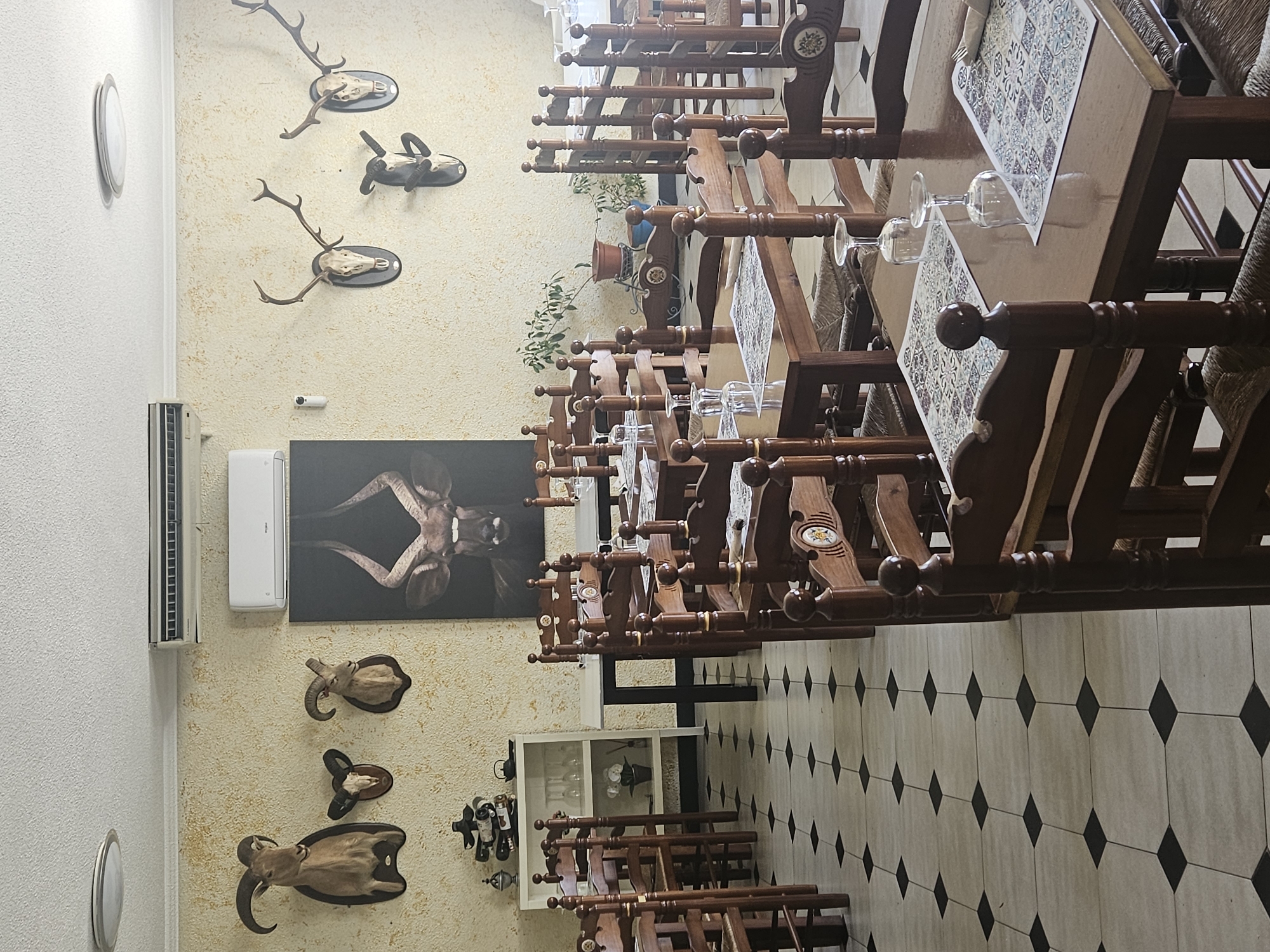 The image depicts the interior of a restaurant or dining establishment. The floor is tiled with a white and black diamond pattern. Several dark wood tables are visible, each set with glassware and decorative table coverings. Matching wooden chairs are positioned around the tables. The walls are beige with a textured finish and adorned with taxidermied animal heads and antlers, along with a framed print of an animal. An air conditioning unit is mounted on the wall. In the background, a white cabinet displays bottles and glassware. The scene suggests a rustic or hunting-themed restaurant. The location is not apparent in the image but presumed to be Los Caballos, Spain.