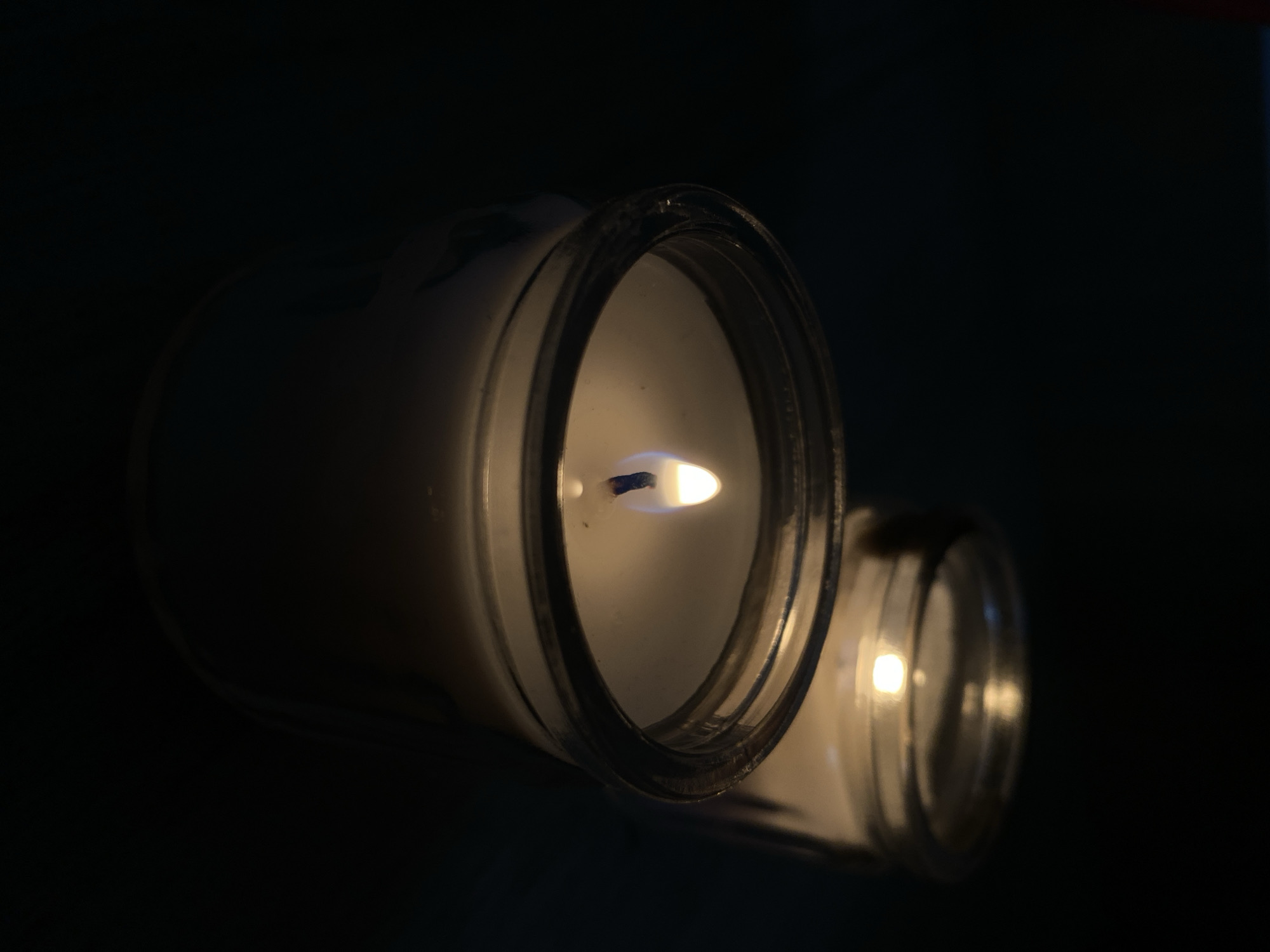 The image features two lit candles, each contained within a clear glass jar, set against a predominantly dark, undefined background. The candle in the foreground is sharply in focus and is oriented horizontally, showcasing its white wax and a vivid flame. The flame displays a distinct blue core at its base, transitioning into bright yellow and orange hues as it extends upward. The rim of this glass jar is visible around the top edge of the candle.
A second candle, also lit and housed in a glass jar, is positioned in the background to the right, appearing blurred and out of focus, contributing to the depth of the scene. Its light is a softer, diffused glow compared to the sharp flame of the foreground candle. The dark environment suggests the photo was taken at night or in a room with minimal ambient light, emphasizing the candles as the sole or primary light source. The scene evokes a sense of calm and warmth. No text is visible in the image.