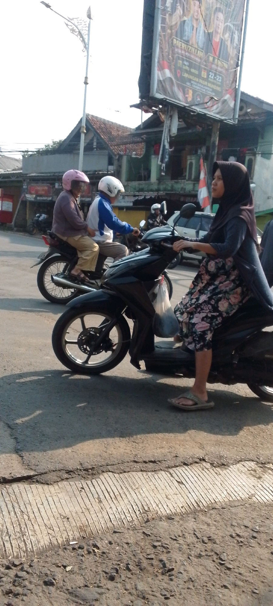 A low-angle, vertical, full shot captures a street scene in Cilaku, Indonesia, bathed in bright sunlight. In the foreground, a black motorcycle is seen from the side, its front wheel and part of its chassis visible. A woman wearing a dark hijab and a floral-patterned skirt is seated on the motorcycle, holding the handlebars. Her right foot, clad in a grey flip-flop, rests on the ground. To her left, another motorcycle is partially visible, carrying two individuals. The rider, wearing a white helmet, is obscured, while the passenger, a person with a pink helmet, is seated behind the rider. This second motorcycle is positioned slightly behind and to the left of the foreground motorcycle. Further in the background, another motorcycle with a rider wearing a white helmet is visible, alongside a white car. Several buildings with tiled roofs line the street, and a tall street lamp with an ornate, decorative top stands prominently on the left side of the image. A large billboard with images of people and text in Indonesian is mounted on a pole in the upper right quadrant of the frame. The road surface is a mix of asphalt and textured concrete, showing signs of wear and tear.