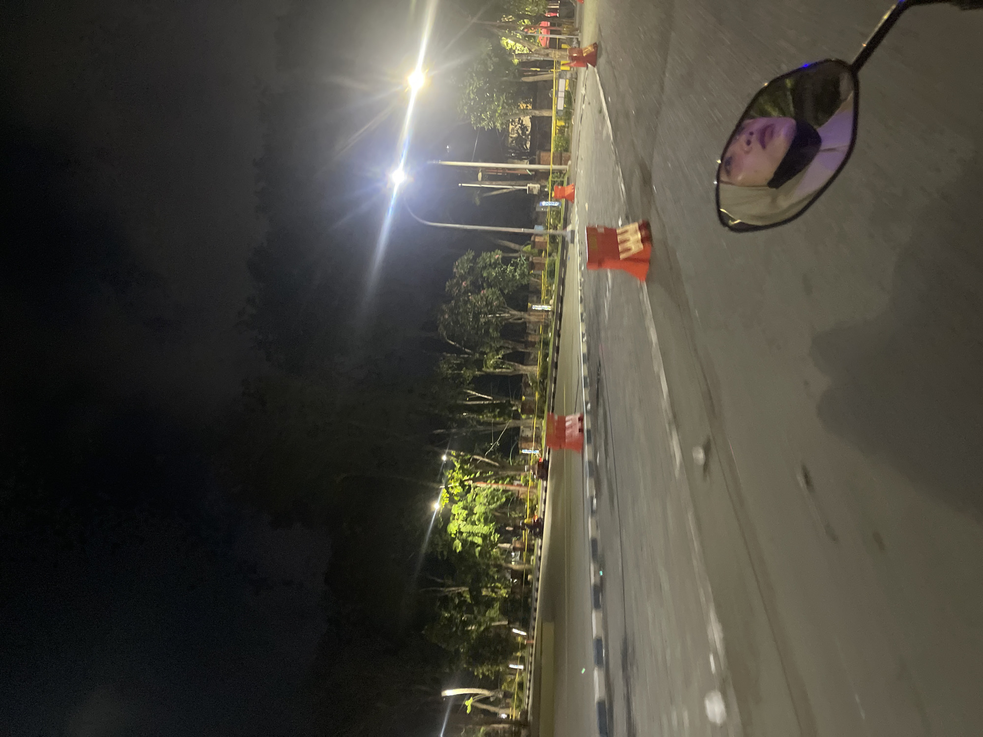 The image depicts a night scene on a road in Sidoarjo, Indonesia. The view is from the perspective of someone riding a motorcycle, as evidenced by the motorcycle's side mirror reflecting a person's face. The person in the mirror is wearing a headscarf and a black face mask. The road appears to be a multi-lane street with streetlights illuminating the scene. Orange traffic barriers are placed along the roadside, suggesting ongoing construction or road maintenance. Trees line the side of the road, their foliage illuminated by the artificial lights. In the distance, more streetlights and some blurred figures, possibly other vehicles or people, can be seen. The overall atmosphere is dark, with the lighting creating stark contrasts and a somewhat grainy appearance due to the low light conditions.