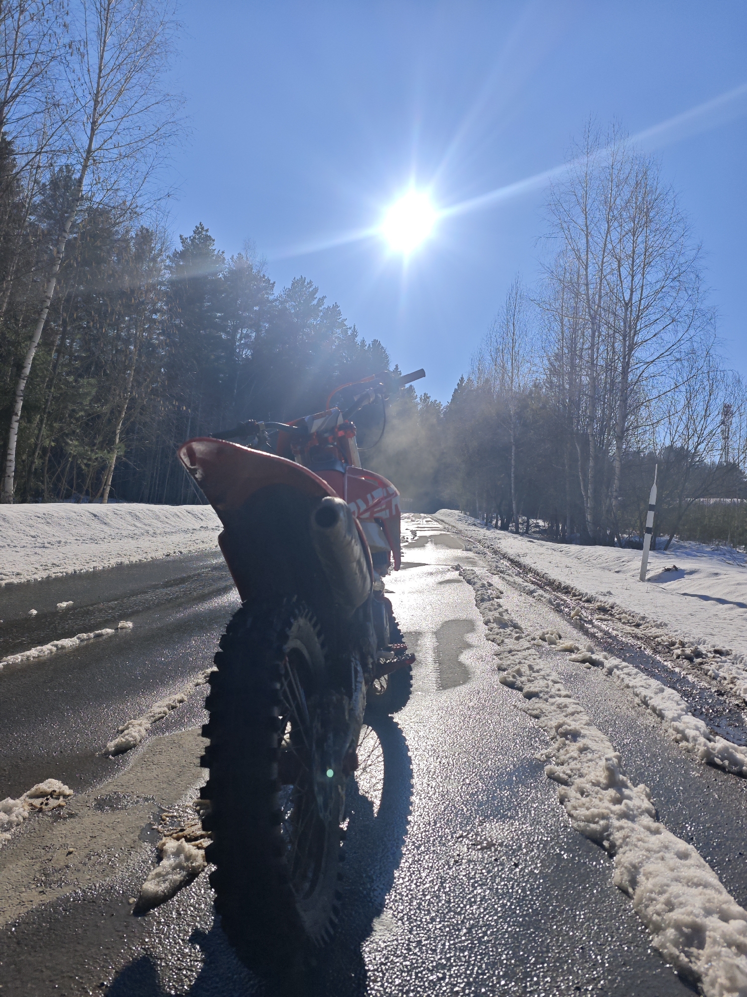 The image depicts a red dirt bike parked on a paved road, partially covered with melting snow and ice, in Прикамский, Russia. The road stretches into the distance, flanked by snowy verges and a forest on both sides. The sun is high in the clear blue sky, creating glare and backlighting the motorcycle. Steam or exhaust is visible near the middle of the bike. A road marker is seen to the right side of the road.