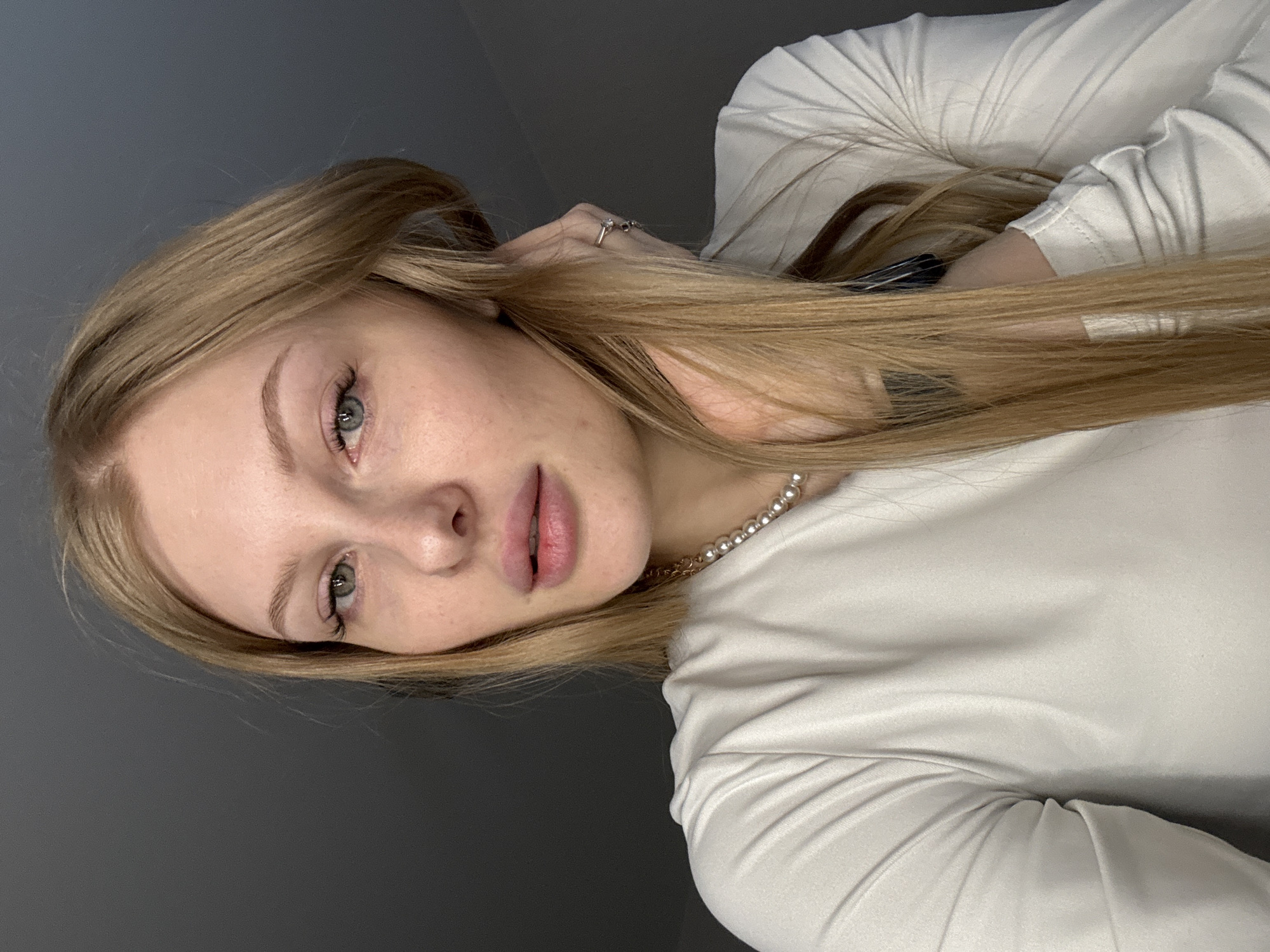 The media file is a close-up portrait of a young woman, presented rotated 90 degrees clockwise from an upright position. She has long, straight, light blonde hair that falls over her shoulders and upper body. Her fair skin shows natural texture and some minor blemishes or freckles. She has light-colored eyes, appearing green or hazel, and well-defined eyebrows. Her lips are naturally full, light pink, and slightly parted.
The woman is wearing a simple, light-colored (off-white or cream) long-sleeved top with soft fabric folds. Around her neck, she wears a pearl necklace with a gold chain. A silver-toned ring with a dark stone is visible on the ring finger of her right hand, which is positioned near her ear, partially covered by her hair. A dark hair clip is also partially visible in her hair closer to her right shoulder.
She is looking directly at the viewer with a calm and neutral expression. The background is a plain, dark gray wall, indicating an indoor setting, possibly a studio, with soft and even lighting. There are no other objects or environmental details visible. No text is present in the image. The image itself provides no visual cues regarding its geographical location.