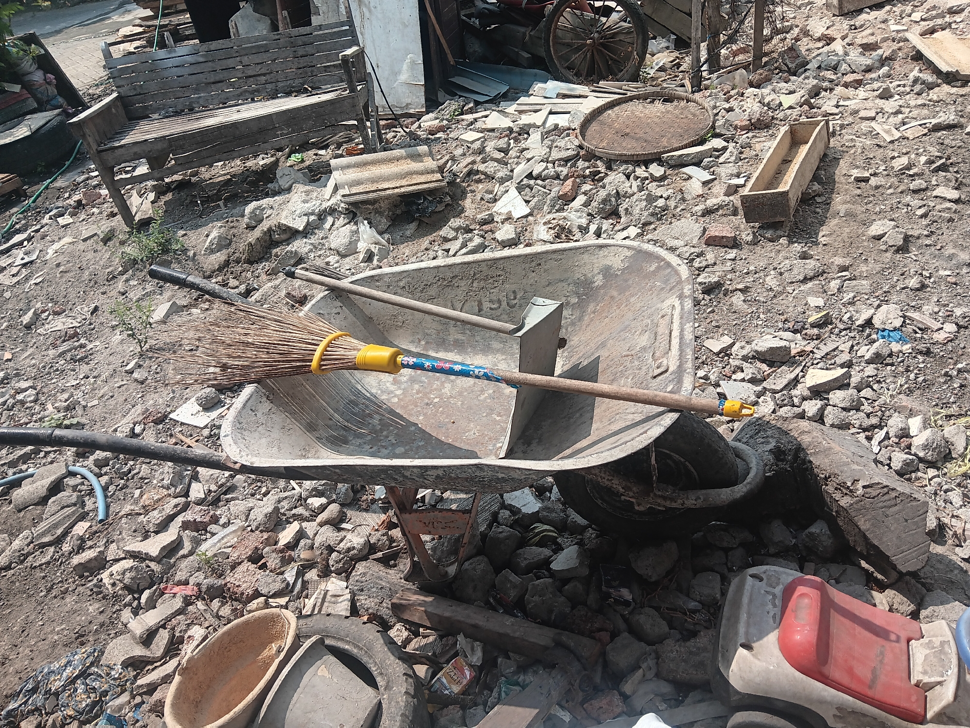 The image depicts a cluttered outdoor scene, likely a construction or demolition site in Batursari, Indonesia. In the foreground, a large metal wheelbarrow is prominent, its basin filled with debris and a broom with a yellow handle and a blue and floral-patterned shaft lying across it. To the left of the wheelbarrow, a black hose and a blue pipe snake across the ground. Behind the wheelbarrow, a weathered wooden bench is situated on a pile of rubble. The ground is covered in a mixture of dirt, broken concrete, rocks, and various discarded items, including what appears to be roof tiles and a wooden trough. In the background, partially obscured, are wooden structures and a spoked wheel. The overall impression is one of disarray and the remnants of a building project or cleanup effort. No people are visible in the frame.