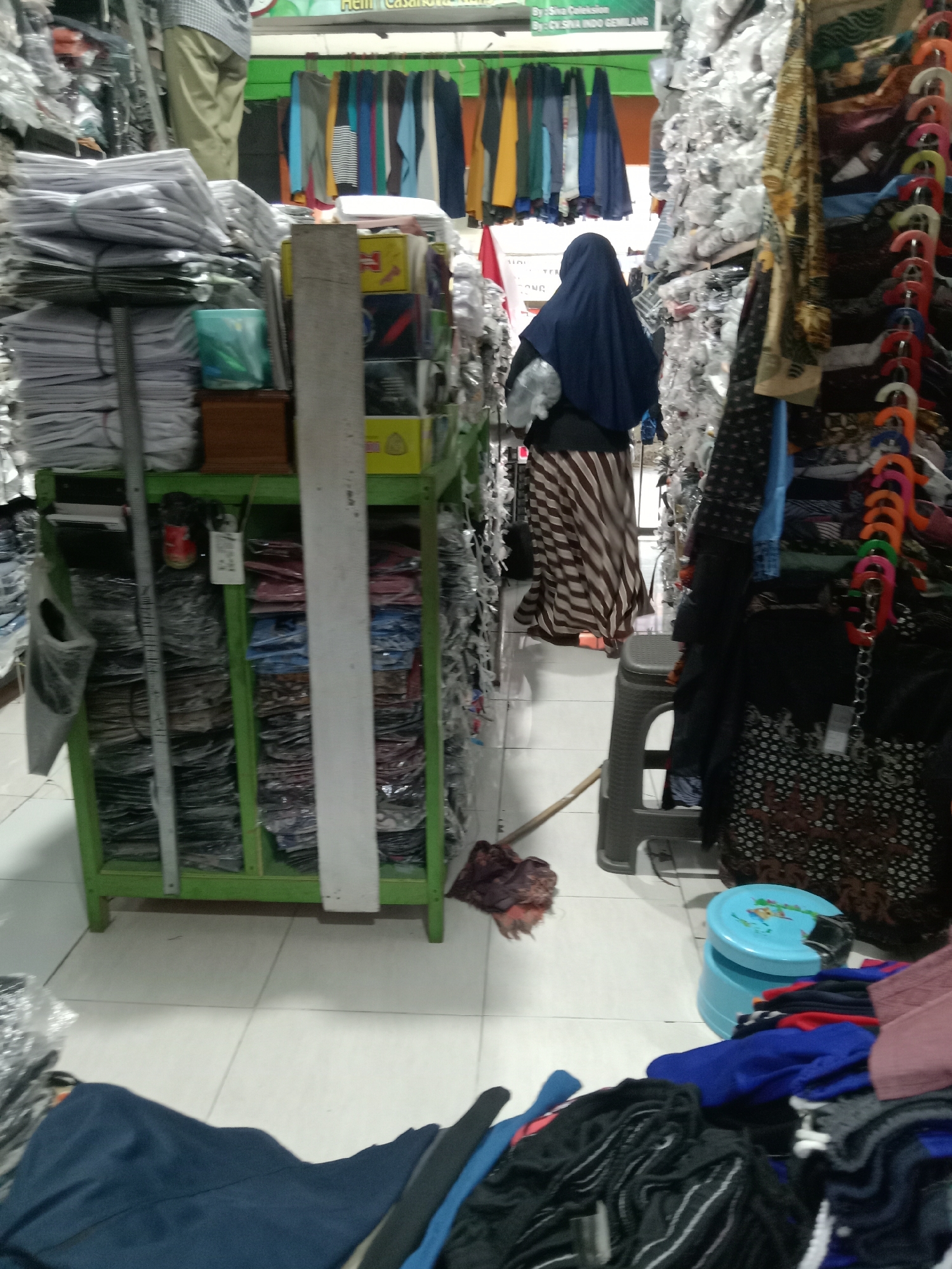The image shows the interior of a clothing store in Sugihwaras, Indonesia. In the foreground, piles of dark clothing are visible on the tiled floor. To the right, a person wearing a dark blue hijab and a striped skirt is standing. Next to them is a gray plastic stool. A blue circular stool with a design on it is also present, along with more piles of clothing in various colors. Further to the left, a green metal shelving unit is filled with stacks of folded clothes in plastic packaging. Behind the shelves, clothing items are hanging on racks, with some appearing to be shirts in different colors. A broom with a red and dark cloth head is lying on the floor near the stool. The overall environment appears to be a retail space with goods displayed for sale.