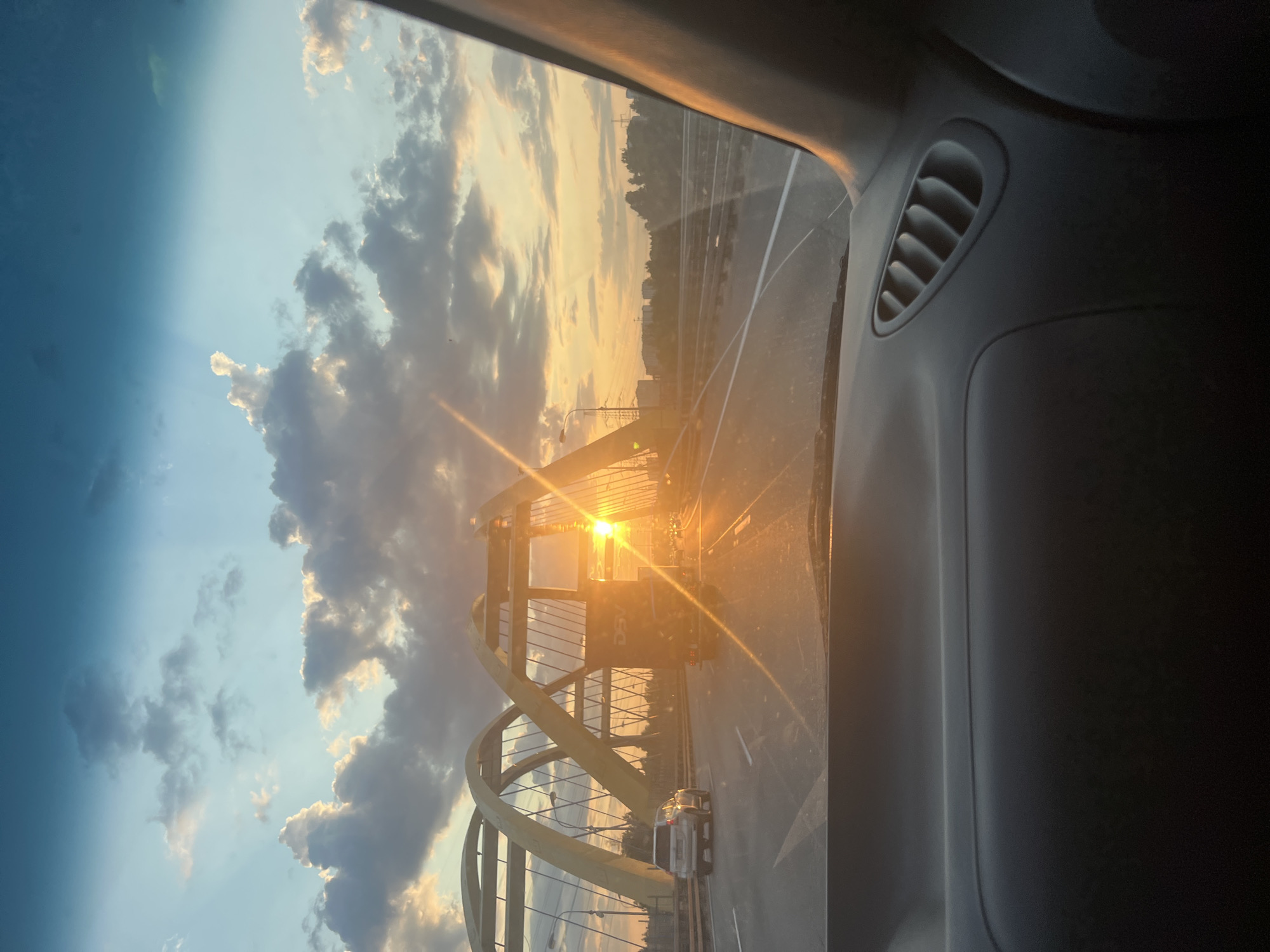 The image captures a view from inside a vehicle, looking forward towards a distinctive yellow arch bridge during sunset. The sun is low on the horizon, casting a warm golden glow and creating lens flare across the scene. Several vehicles are visible on the multi-lane highway, including a silver SUV in the left lane and a large truck with "DSV" lettering on its side in the right lane. The truck is directly in front of the sun, obscuring it partially. The bridge's structure, composed of large yellow arches and supporting cables, dominates the mid-ground. In the distance, a city skyline with buildings and trees can be seen silhouetted against the colorful sky. The sky itself is a gradient of blues and oranges, with scattered clouds illuminated by the setting sun. The foreground shows the interior of the car, with the dashboard and a ventilation grille clearly visible. The scene appears to be on a highway, possibly crossing the Neva River in Saint Petersburg, Russia, given the presence of a large bridge structure and a city skyline.