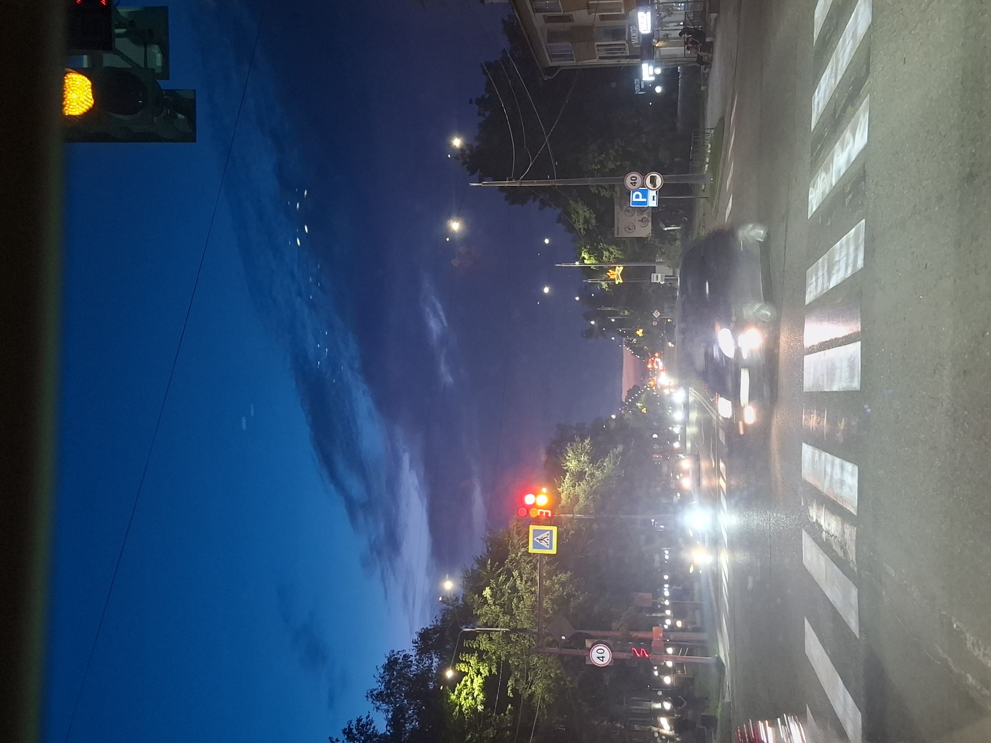 The image depicts a street scene at dusk or night in the city of Svobodny, Russia. A traffic light is visible, showing a red and yellow signal. The street is wet, indicating recent rain or it's a damp evening, and the road markings for a pedestrian crossing are clearly visible. Several cars are on the road, with headlights illuminating the scene. Some trees line the street, and buildings are visible on the right side. The sky is a deep blue, with some clouds and distant lights. The overall impression is of a city street after dark, with traffic and pedestrian activity.
