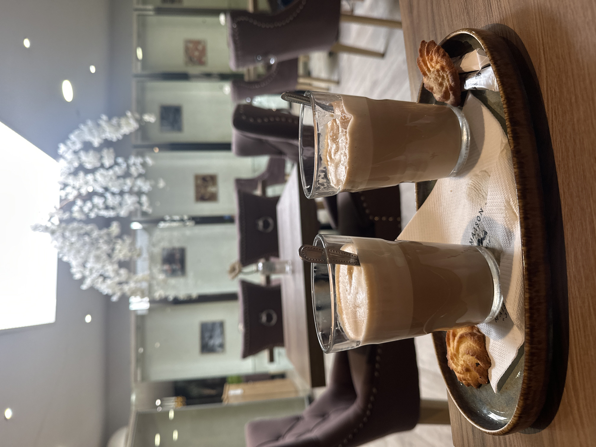 Two glasses of coffee with foam are on a tray with two cookies and two sugar packets. The tray is on a wooden table in a cafe. The cafe has plush purple chairs and tables. There are white floral decorations hanging from the ceiling. The image was taken in Nivelles, Belgium.
