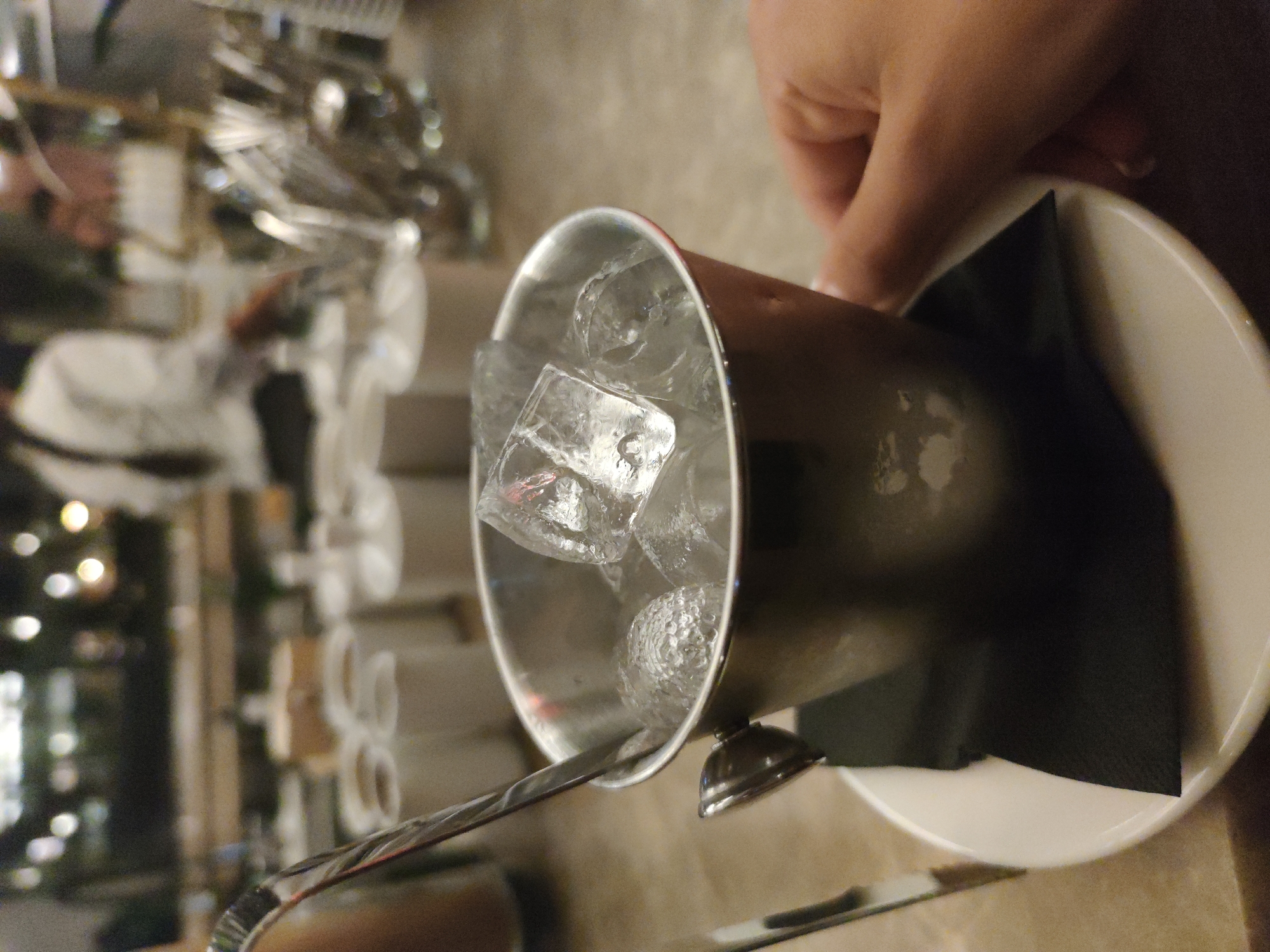 The image captures a close-up of a small, polished metallic bucket filled with clear ice cubes and a metallic scoop, resting on a white ceramic saucer with a folded black napkin underneath. A light-skinned human hand is visible on the right side of the frame, grasping the bucket. The background, significantly blurred, depicts an indoor commercial establishment, likely a restaurant or bar, in the city of Unknown, Unknown. In the mid-ground, a row of white cylindrical containers or cups and a collection of metallic utensils are visible on a counter. The far background reveals indistinct human figures, including a person in a white shirt, and numerous circular ambient light sources, indicating a low-light environment.