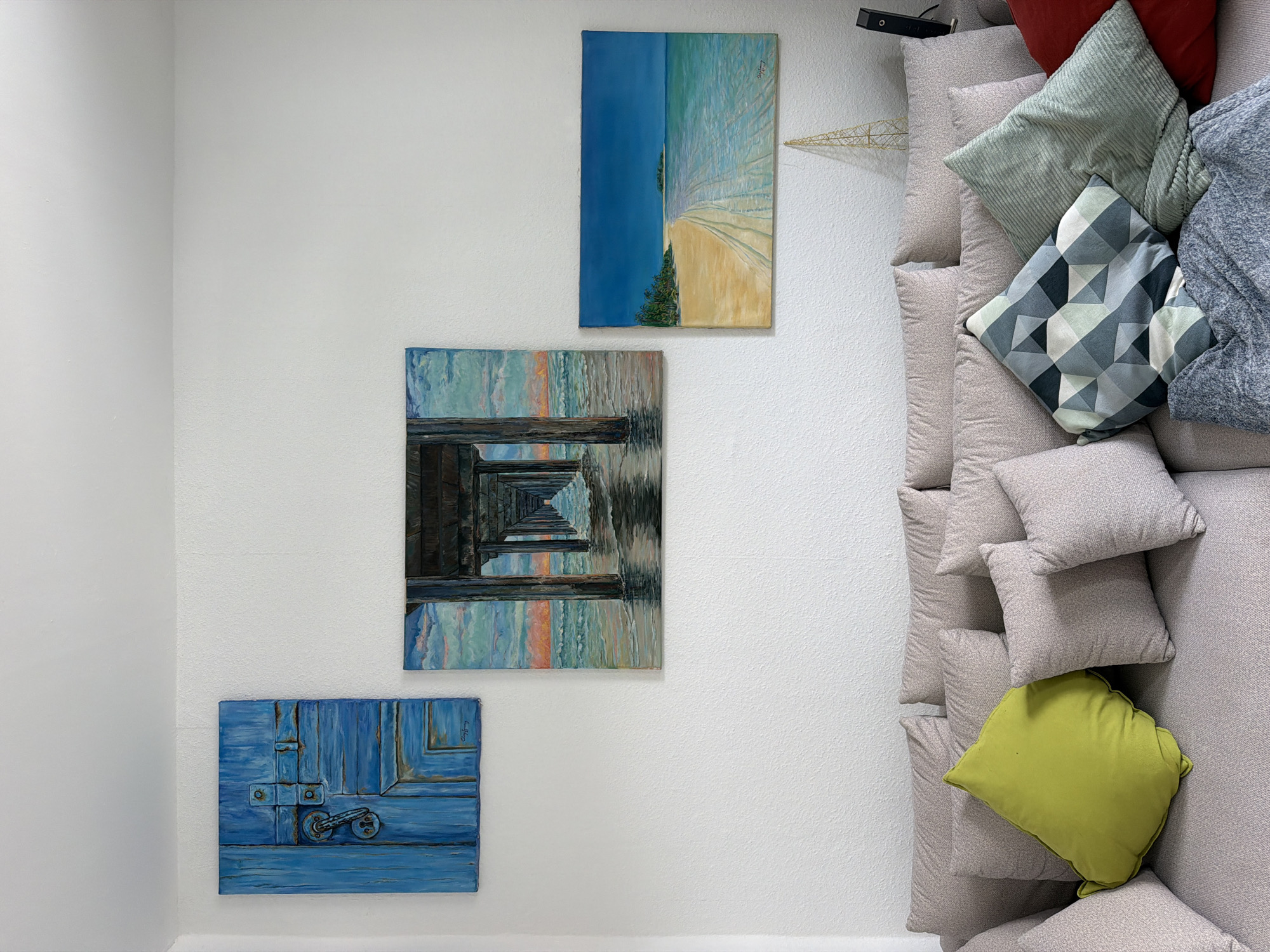 The image shows a living room interior, possibly in Herne, Germany, featuring a neutral-toned sofa adorned with various throw pillows and a blue blanket. Above the sofa, a white wall displays three landscape paintings: a vibrant blue door on the left, a perspective view of a pier with the ocean below in the center, and a tropical beach scene with palm trees and clear blue water on the right. A gold, abstract decorative object and a black electronic device are visible on the right side of the frame. The lighting suggests an indoor setting, likely during the daytime.