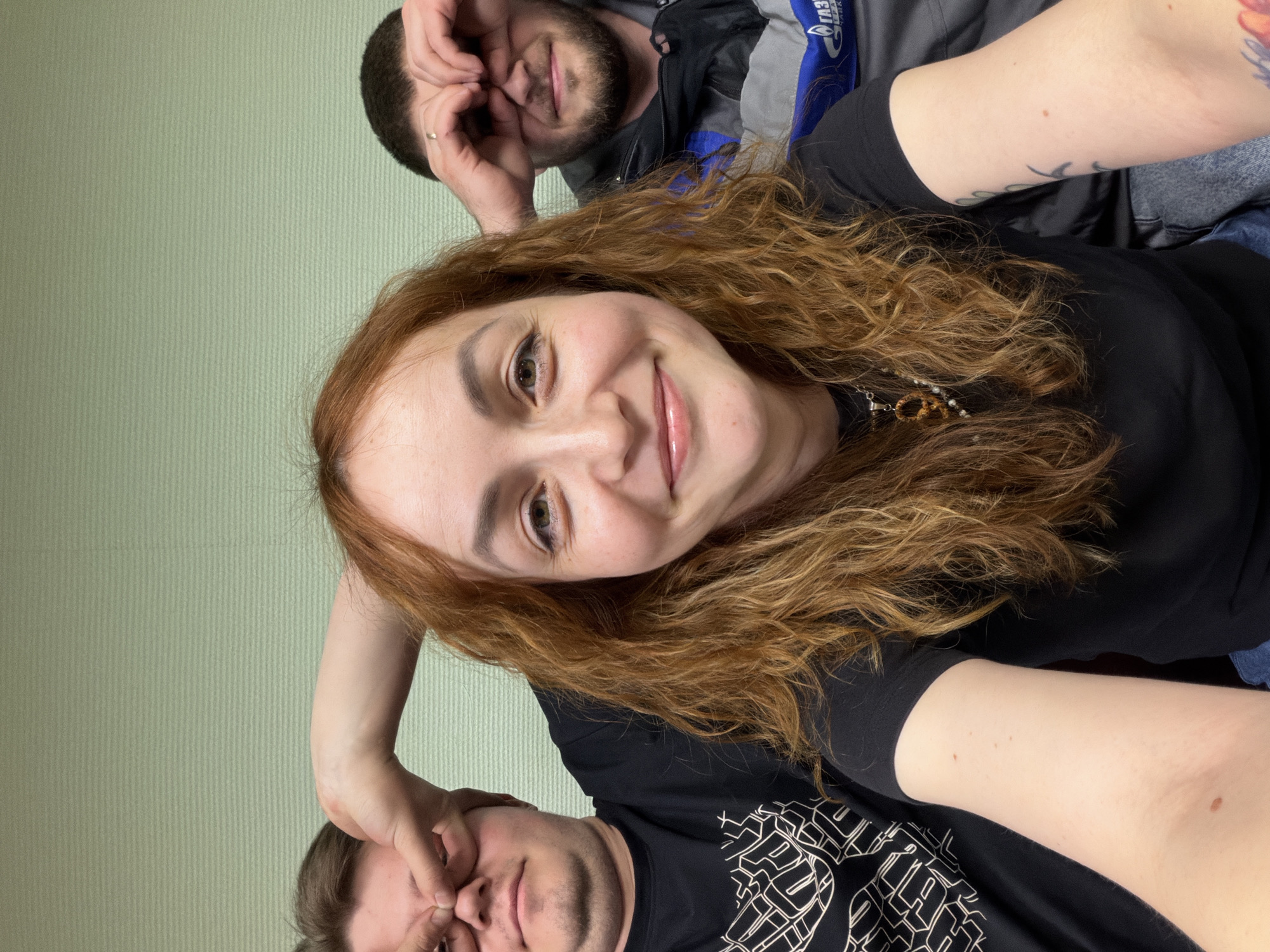 This is a vertical-format selfie featuring three people in an indoor setting, taken in Chaikovsky, Russia. In the foreground, a woman with long, wavy reddish-brown hair smiles warmly at the camera. She wears a black top and a necklace with a pendant and beads, and a tattoo is visible on her right arm.
Behind her, two men are making playful faces. To the woman's left, a man with short brown hair and a beard is pinching his nose and eyes with his fingers. He is wearing a black t-shirt with a white geometric pattern. To the woman's right, another man with short dark hair and a beard forms a "heart" or "glasses" shape with his hands over his eyes. He is wearing a grey and blue jacket with the partial Russian text "ГАЗ" (GAZ) visible, and a ring is on his left ring finger.
The background is a simple, light green wall with subtle vertical lines. The overall mood of the photo is cheerful and casual, suggesting a lighthearted gathering among friends.