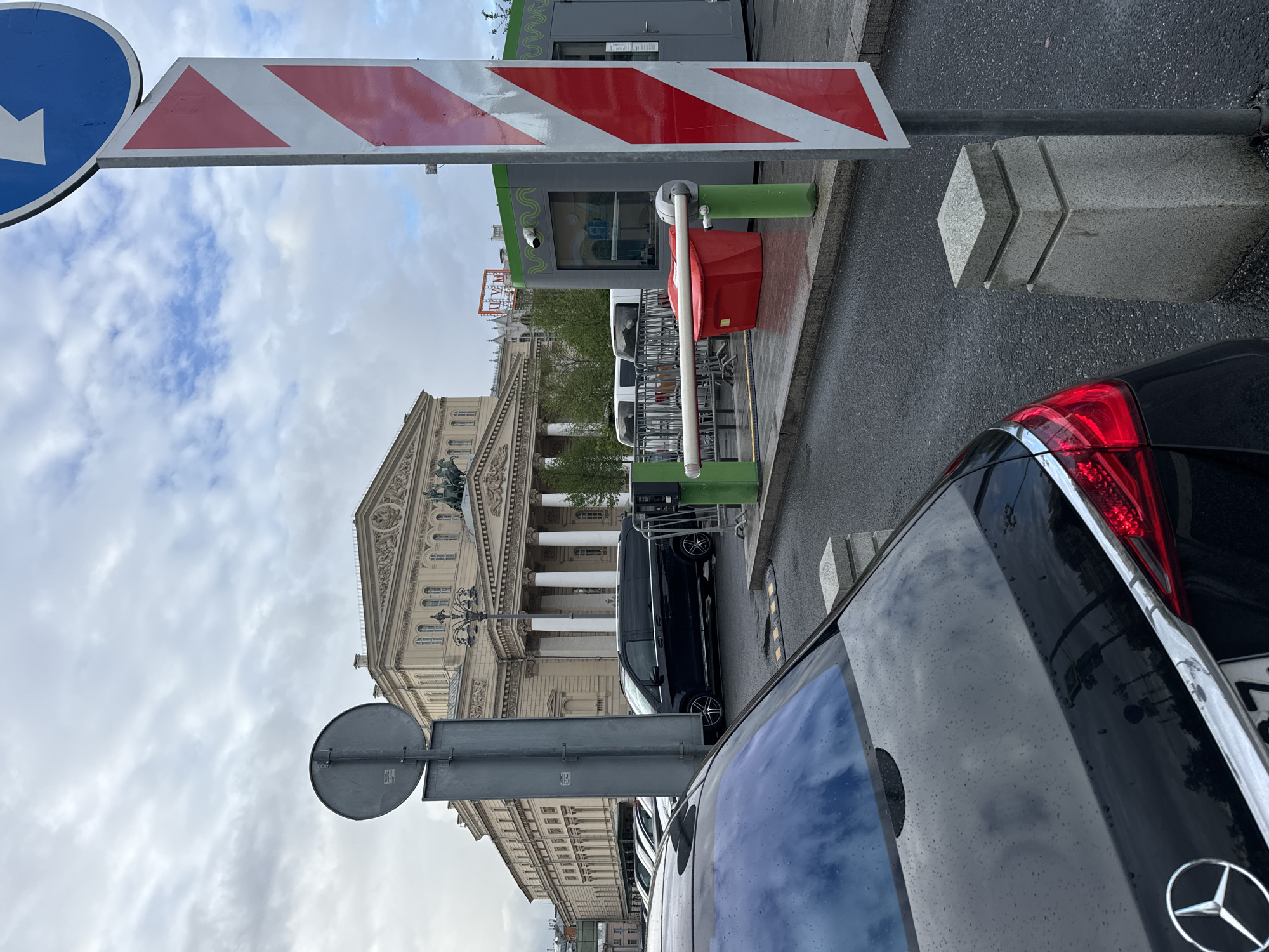 The image depicts a street scene in Moscow, Russia, with the Bolshoi Theatre prominently featured in the background. In the foreground, there's a black Mercedes car parked, showing its rear side with a visible taillight and Mercedes logo. A pedestrian access control point is visible, with a red and white striped barrier and a small booth. Traffic and directional signs are seen on poles. The sky is overcast, and the street appears wet.