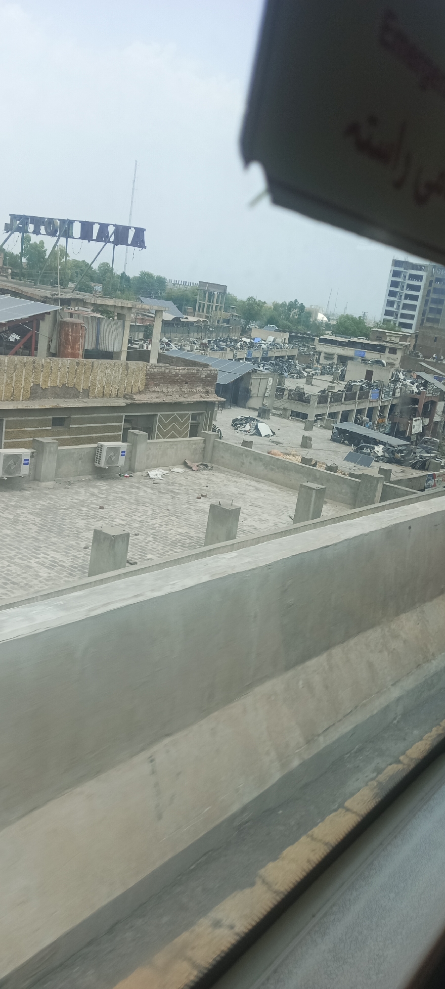The image captures an outdoor scene with a sprawling urban landscape, likely a junkyard or industrial area, under an overcast sky. In the foreground, a concrete barrier with a textured surface runs diagonally across the frame, suggesting the viewpoint is from a raised platform or moving vehicle.
In the midground, a rooftop area is visible, paved with brick tiles and featuring several concrete pillars. Two air conditioning units are mounted on a wall. Beyond this rooftop, the scene opens up to a densely packed area filled with numerous vehicles, many of which appear to be scrap or dismantled. Various structures, including low-rise buildings and what look like temporary shelters with corrugated metal roofs, are interspersed among the vehicles. Some solar panels are also visible on some of the rooftops, indicating a mix of old and modern infrastructure.
Further in the background, a large, industrial-looking structure with a prominent lattice-like framework is visible, along with trees and taller buildings. The overall impression is one of a busy, perhaps somewhat neglected, urban environment. The text "Emergency Exit" is partially visible in the top right corner, written in English and what appears to be Urdu script. The location is identified as Alam Khan Kalay, Pakistan.