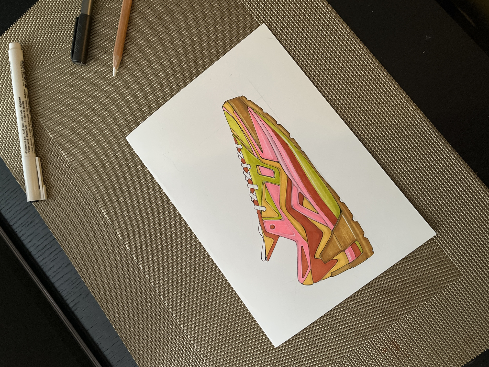 The image shows a drawing of a sneaker on a piece of white paper. The sneaker is colored in shades of brown, orange, pink, and green. Several pens and pencils lie near the paper. The paper and drawing are on a brown, textured placemat. A dark black surface is visible near the lower left and upper right corners of the placemat.