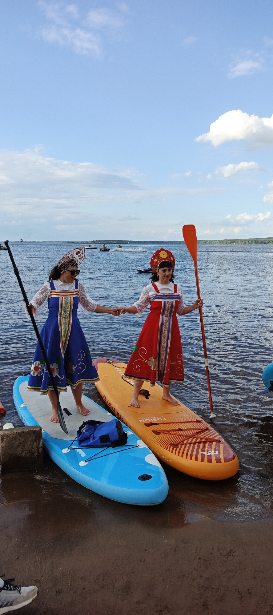 Two women, dressed in traditional red and blue embroidered dresses and head coverings, stand on separate paddleboards in the water. The woman on the left, wearing a blue dress, holds a paddle with her left hand. The woman on the right, wearing a red dress, holds an orange paddle with her right hand. They are holding hands and appear to be interacting.
The paddleboards are positioned next to each other, with the blue board slightly in front of the orange one. A blue dry bag is placed on the blue paddleboard. The background shows a body of water with several boats and jet skis in the distance, and a shoreline with trees. The sky is blue with scattered white clouds. The scene appears to be outdoors, likely on a lake or river, on a clear day.