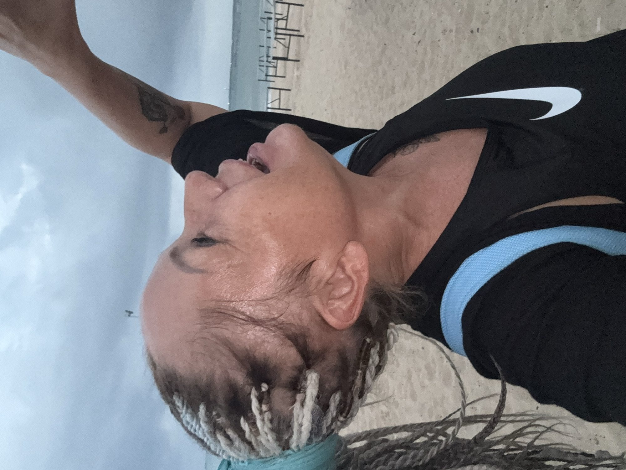 The image shows a close-up, side profile of a woman on a beach. She is wearing a black Nike tank top with a light blue strap visible over her shoulder. Her hair is styled in long braids with some lighter colored sections. She appears to be looking upwards and to the right with her mouth open. Her arm is raised upwards and to the right, with a tattoo visible on her upper arm. The background shows a sandy beach with some dark, metal structures and the ocean and a hazy sky in the distance. The overall lighting suggests it may be overcast or twilight. The image does not contain enough information to confirm the location as Kuta, Indonesia.