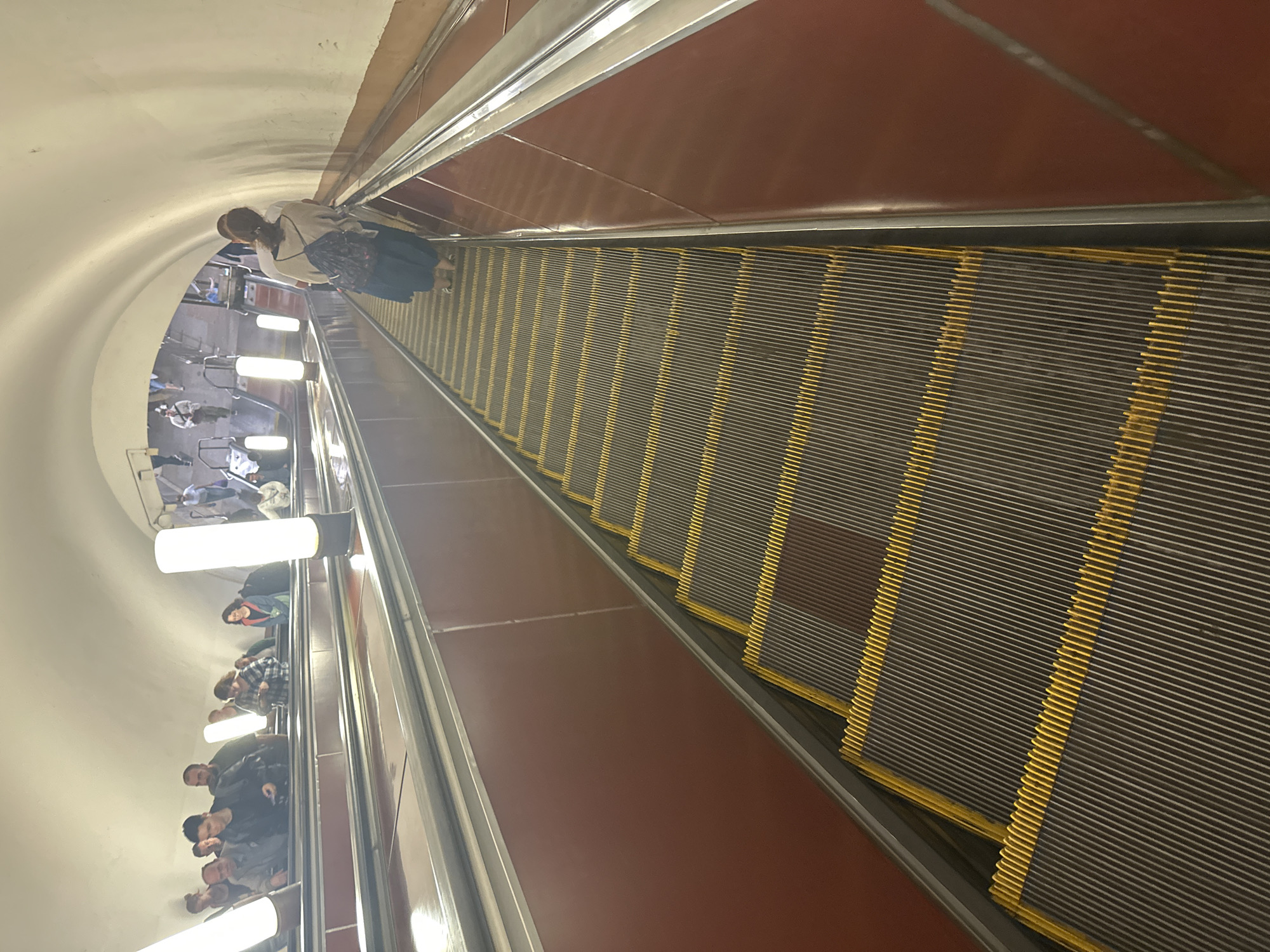 An overhead shot captures a downward-facing escalator in a dimly lit, curved tunnel. Several people are visible on the escalator, some at the top and others further down, with one woman in the foreground wearing a backpack and a light-colored shirt. The escalator steps are dark with yellow safety lines. The tunnel walls are a muted red, and tall, cylindrical lights illuminate the space. The overall atmosphere is utilitarian, suggesting a public transportation hub.