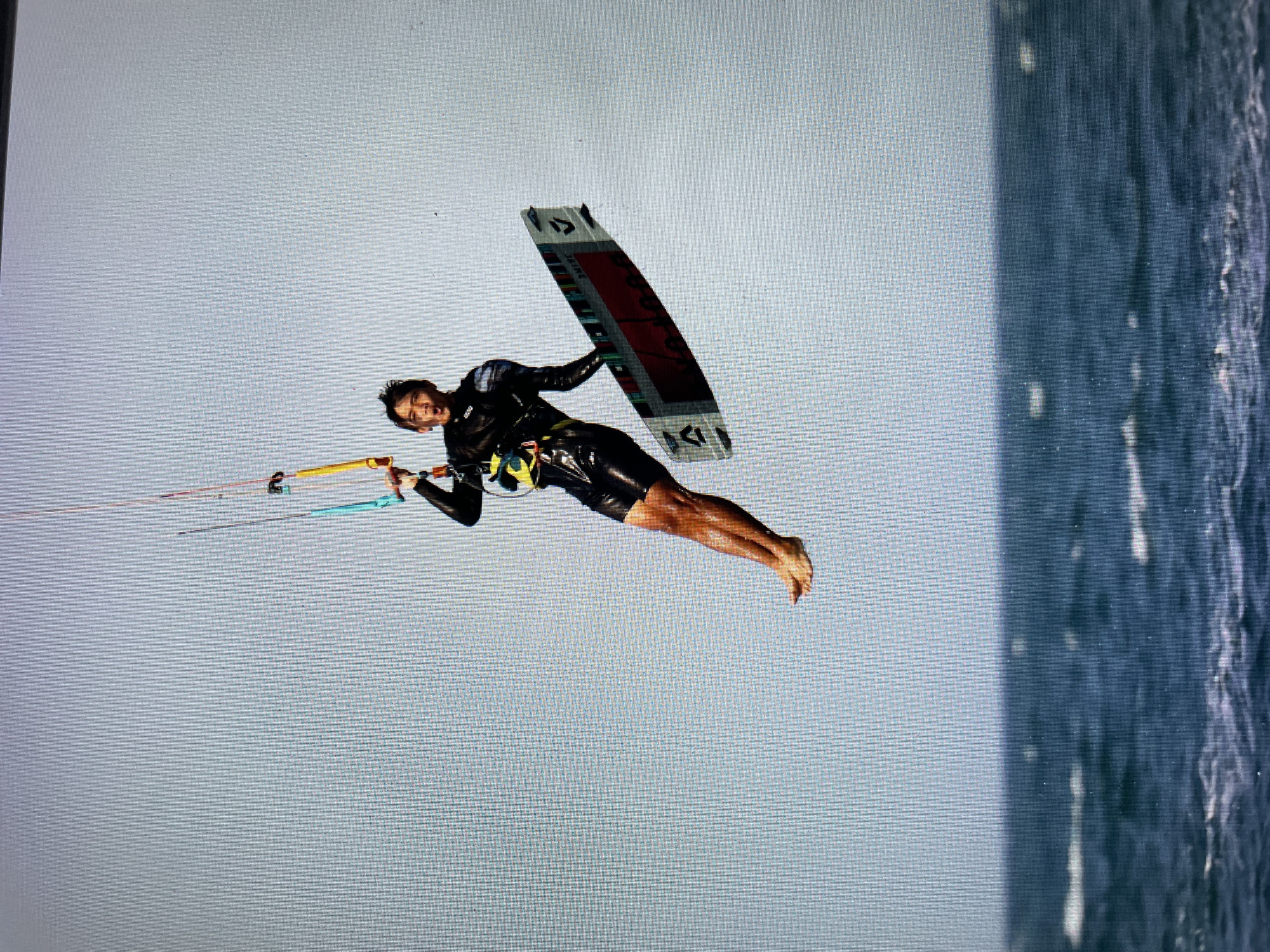 A person in a black wetsuit is kitesurfing in the sea. The location appears to be the coastal area of Bacong, Philippines, with the sea on the right side of the image and a cloudy sky in the background. The individual is airborne, holding a kiteboard with one hand and the kite control bar with the other, executing a trick. The water surface is visible on the right side of the image.