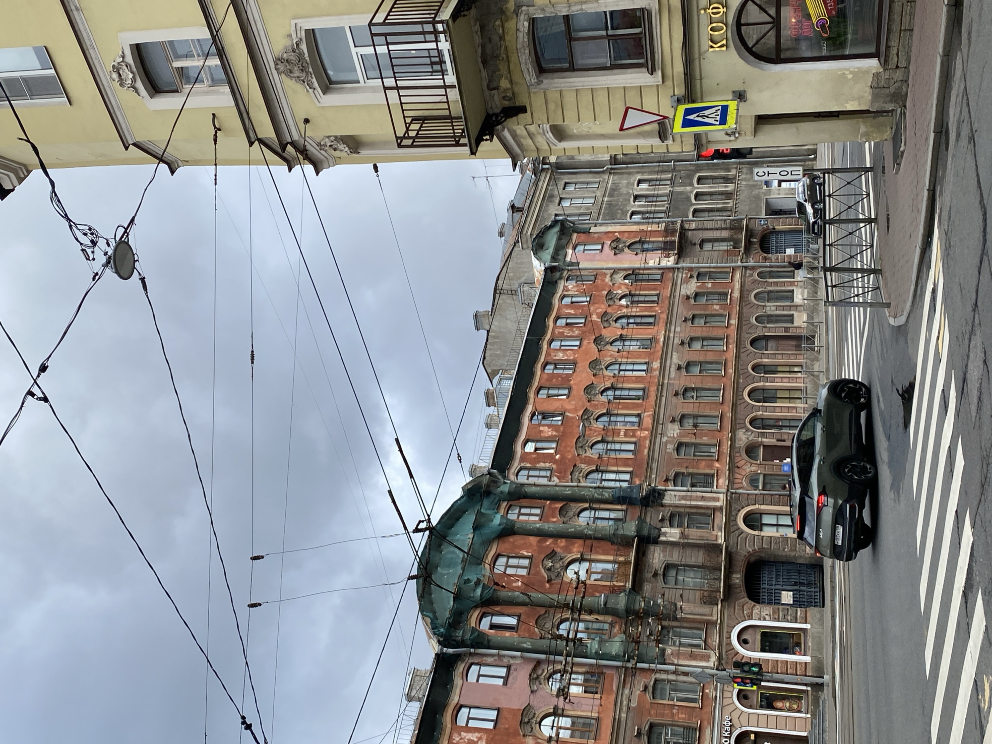 The image depicts a street scene in Saint Petersburg, Russia, on a cloudy day. The foreground features a pedestrian crossing with a dark green SUV driving away from the viewer. To the right, a yellow building with ornate architectural details and a balcony stands at a corner. On this building, a traffic light shows red, and a pedestrian crossing sign is visible. Further down the street, several older buildings with colorful facades and numerous windows are lined up. Overhead, a complex network of tram wires crisscrosses the sky. The architecture suggests a historical urban setting.