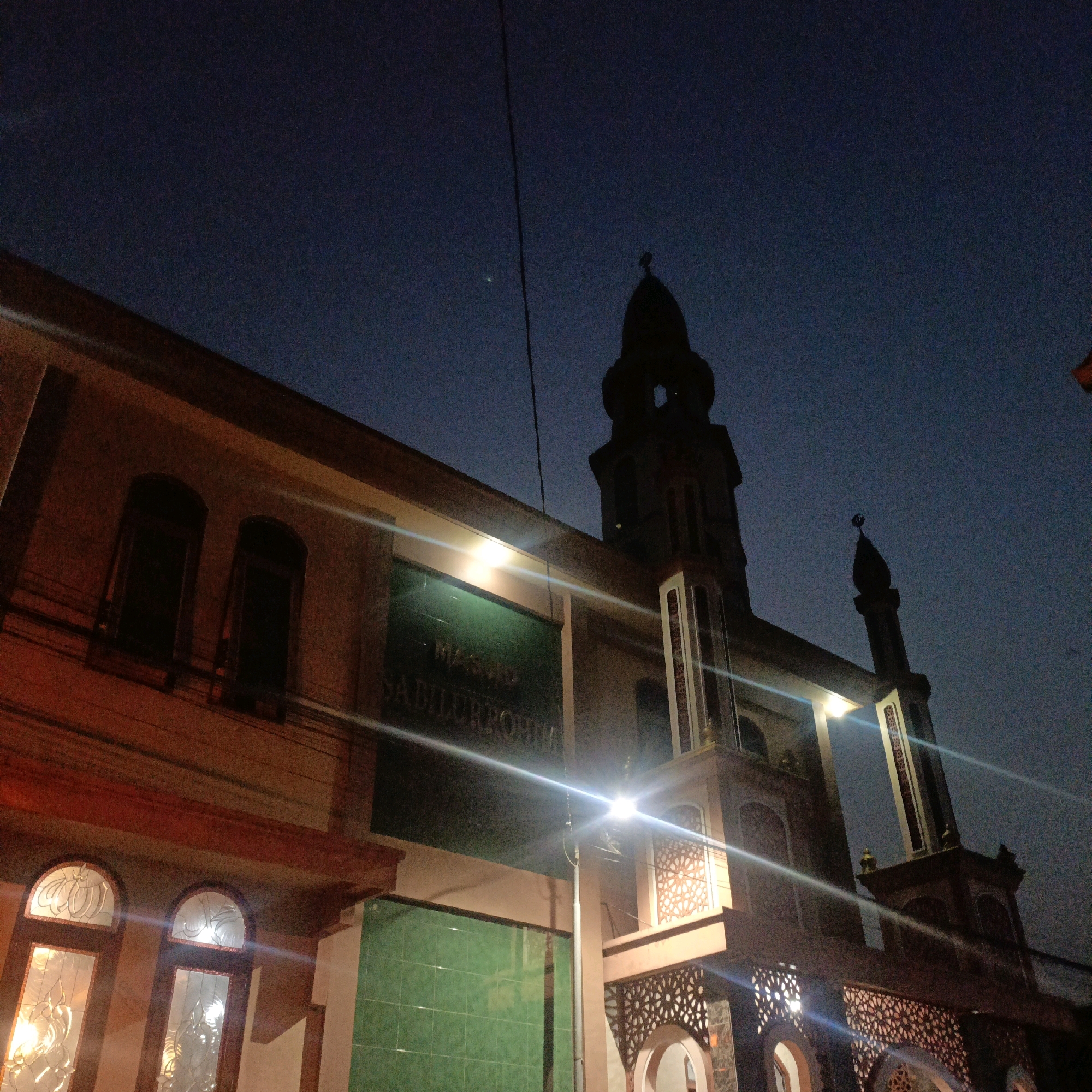 The image shows the exterior of the MASJID SADILLURROHMAN mosque in Salatiga, Indonesia, at dusk. The mosque features a tall, slender minaret topped with a crescent moon and star, characteristic of Islamic architecture. The building's facade is illuminated by interior lights, casting a warm glow on the windows and architectural details. Ornate latticework adorns the entrances and balconies, adding a decorative touch. The sky is a deep blue, transitioning into darkness, suggesting the end of daylight. The overall scene captures the mosque's presence in its urban environment.