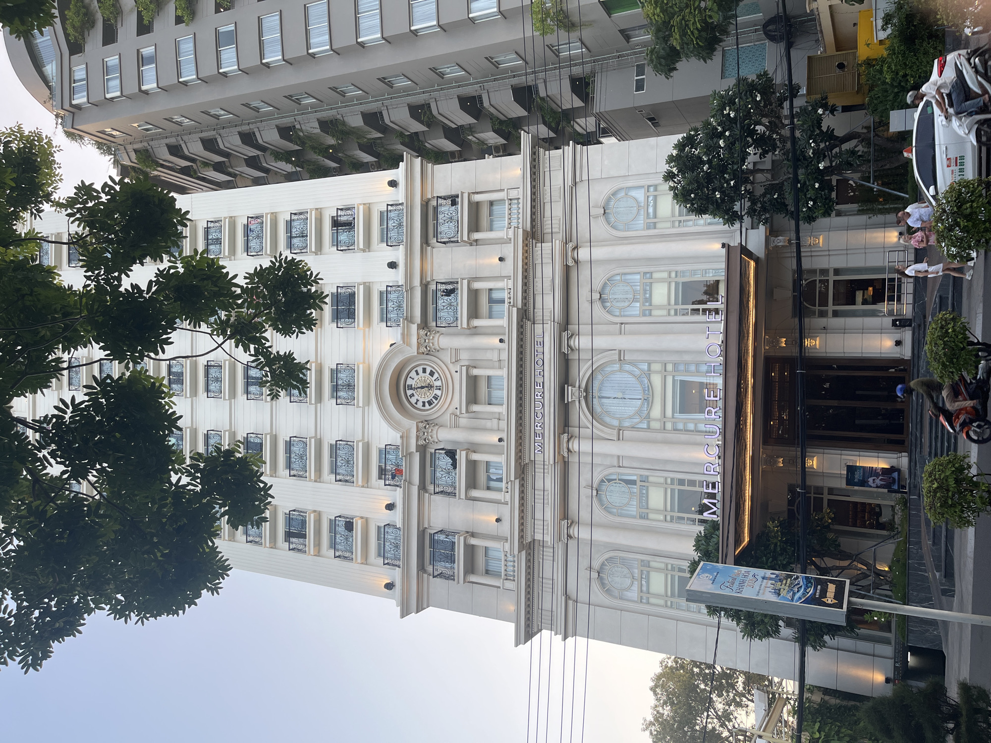 The image shows the Mercure Hotel in Nha Trang, Vietnam. The hotel is a tall, white building with many floors, each featuring balconies with black railings. A large clock is visible on the facade of the building. In front of the hotel, there is a street with traffic, including a motorcycle with a rider and a car. Several trees and bushes are present, some with white flowers. People are seen standing and walking near the entrance of the hotel. The sky is clear and a bit hazy, suggesting it might be morning or late afternoon. The architecture of the Mercure Hotel is elegant, with classical columns and arched windows on the lower floors. The adjacent building is a gray high-rise with balconies and greenery.