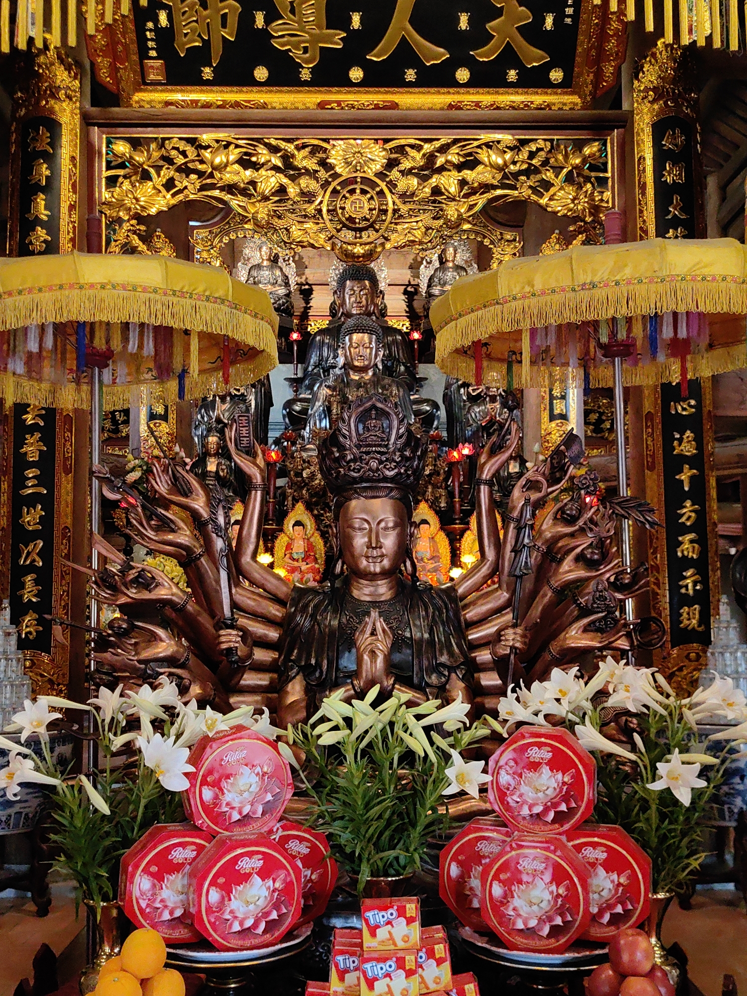 This image depicts a Buddhist shrine in Hong Gai Ward, Vietnam. The central figure is a bronze statue of a seated Buddha with multiple arms, each holding different objects. Above and behind this statue are two more Buddha figures. The shrine is adorned with intricate gold carvings and yellow tasseled umbrellas. Offerings of white lilies, red circular boxes with floral designs, oranges, apples, and boxes of Tipo biscuits are placed in front of the statue. Inscribed vertical banners flank the shrine.