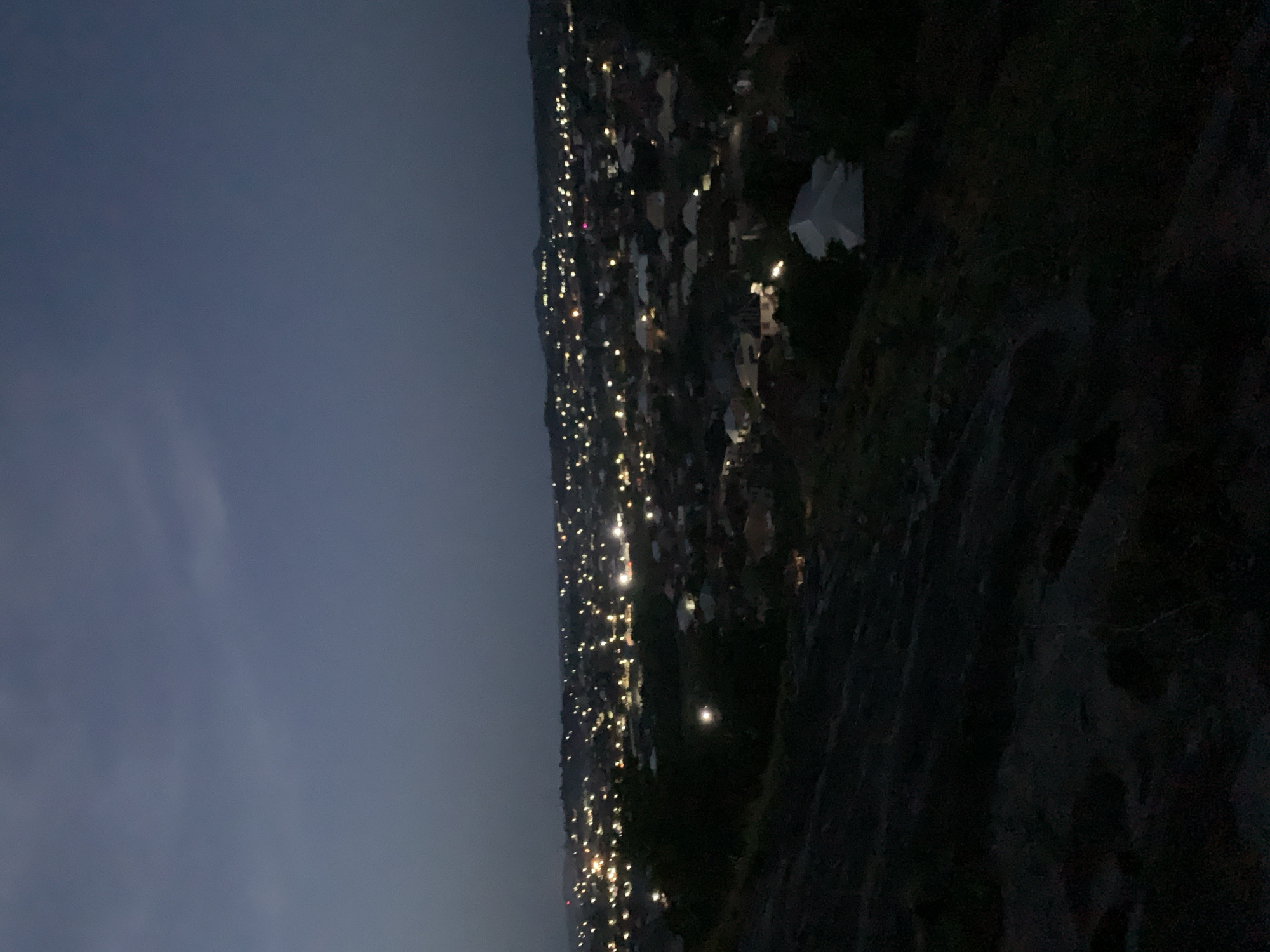 At night, a view overlooks the city of Kabba, Nigeria, where numerous lights from houses and buildings illuminate the landscape. The sky is a dark blue, transitioning to a lighter shade near the horizon. In the foreground, the dark, textured ground slopes downward.