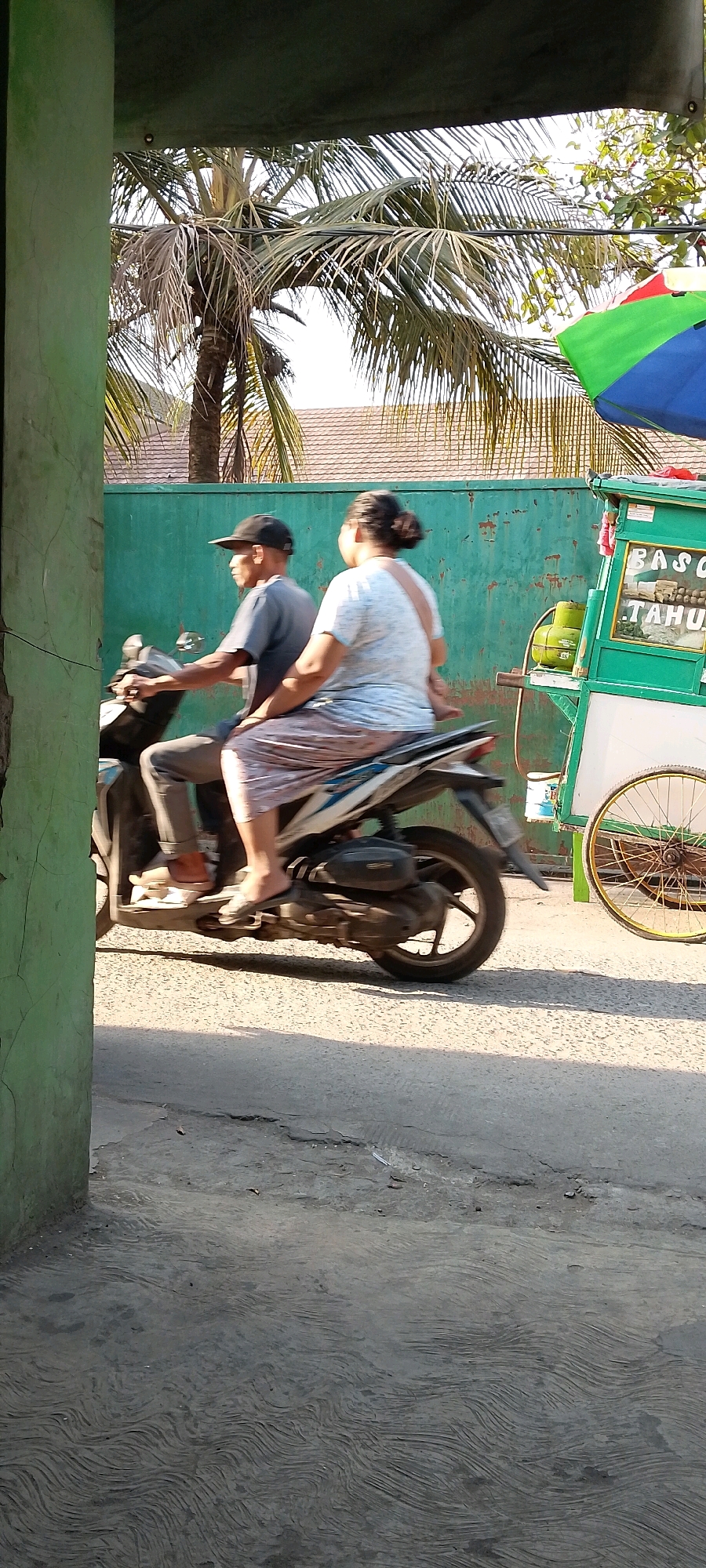 In Karangrahayu, Indonesia, a man wearing a black cap and a gray t-shirt rides a motorbike. A woman with her hair pulled back in a bun, wearing a light blue t-shirt and a patterned skirt, is seated behind him. They are moving past a green wall, with a palm tree visible in the background. To the right, a green food cart is visible, with a propane tank and a yellow-wheeled bicycle attachment. The cart displays the words "BASO TAHOE" and appears to be a food vendor. The scene is illuminated by sunlight, casting shadows on the asphalt road.