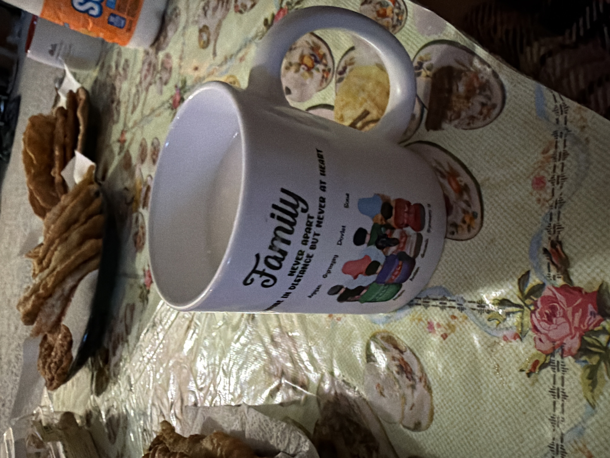 The image depicts a close-up, slightly tilted view of a table covered with a light green, patterned tablecloth. On the table are several food items, including what appear to be fried pastries or dough sticks arranged on a dark plate. A white mug is prominently featured, with a message and illustrations on its side. The mug contains a light-colored liquid. To the right of the mug, a portion of a white bottle with orange and blue lettering is visible. The background is indistinct but suggests an indoor setting. The overall impression is of a casual meal or snack setting. The specific location in Kirkland, United States of America is not discernible from the image itself.