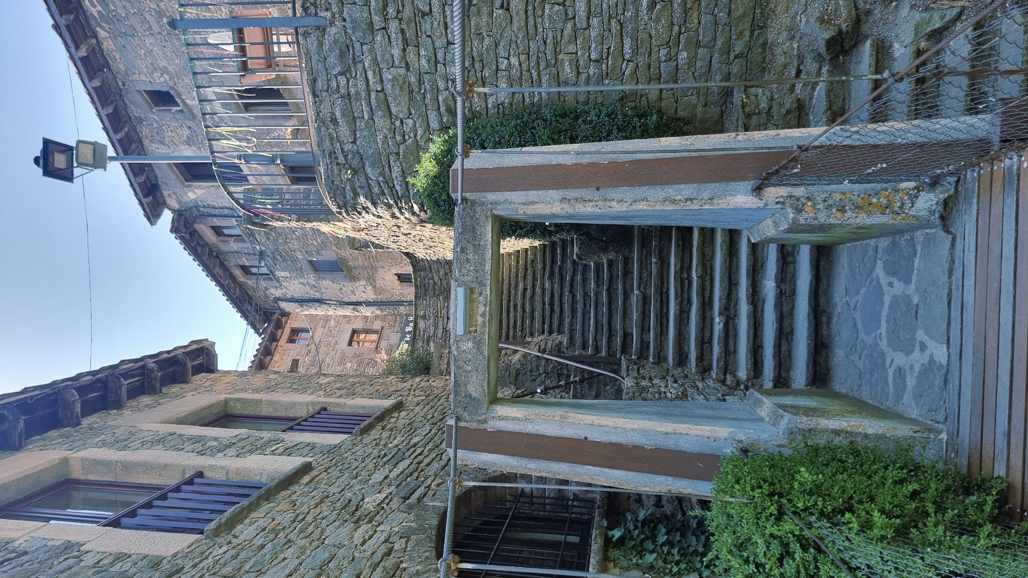The image depicts a narrow stone stairway in the historic village of Rupit i Pruit, Spain, likely in its medieval quarter. The stairway is framed by rough stone buildings with slate roofs, characteristic of the region. A rustic stone archway marks the entrance to the stairs. The steps themselves are made of stone, and the path at the bottom appears to be paved with flagstones. The scene is bathed in natural sunlight, casting shadows and highlighting the textures of the stone. Greenery, including ivy on the left building and a dense shrub to the right of the stairs, adds a touch of nature to the stone architecture. A metal railing and some wire fencing are visible along the sides of the path and stairs, suggesting a functional or safety feature. A street lamp stands on a pole above the buildings on the right. There are no people or animals visible in the frame.