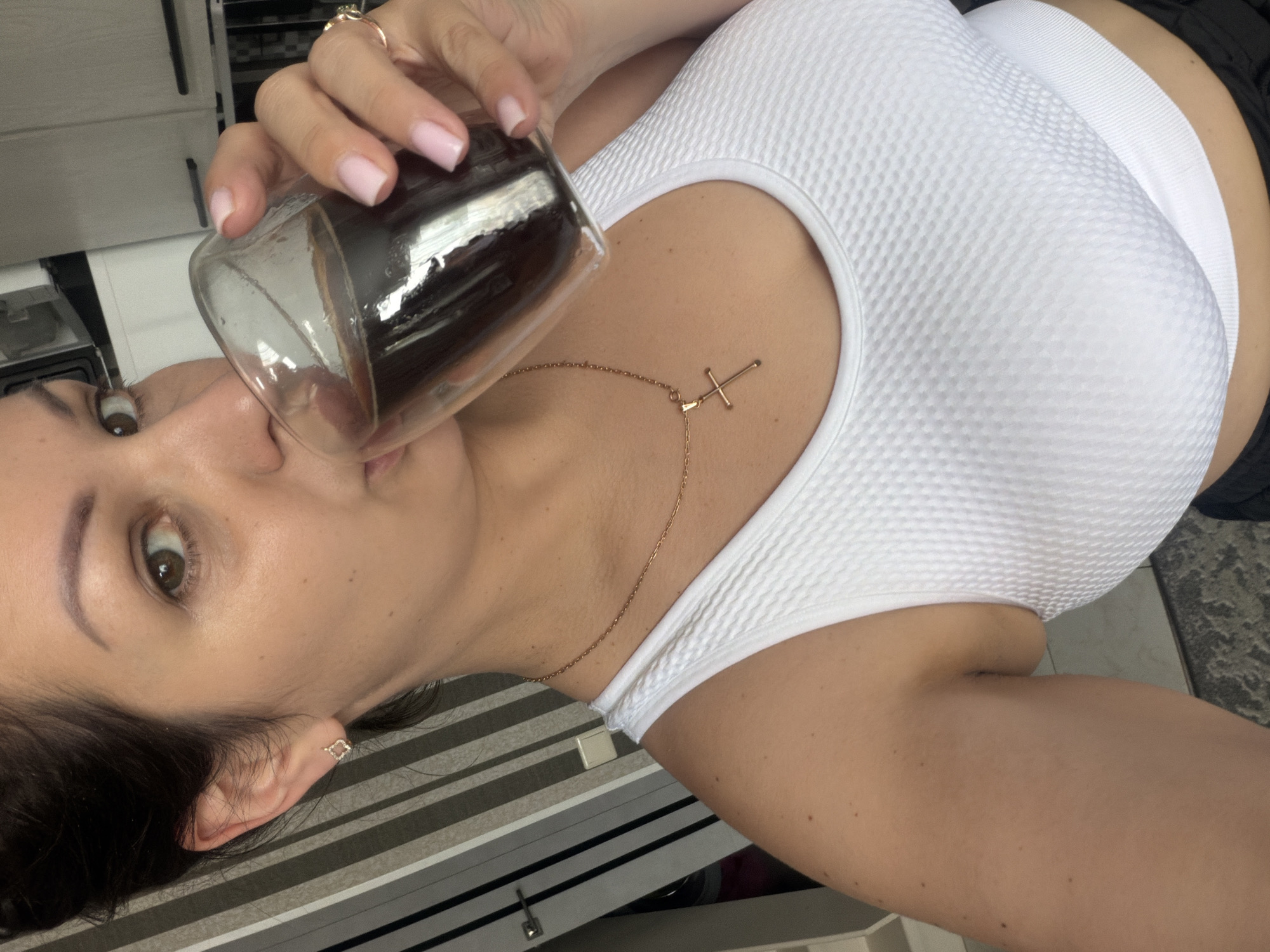 A woman with dark hair pulled back is taking a sip of coffee from a clear glass. She is wearing a white sports bra and a gold necklace with a cross pendant. She has a diamond stud earring in her left ear and a ring on her right hand. The background appears to be a kitchen with striped wallpaper and cabinets. The image is a close-up of the woman's face and upper body. The image is not in Sochi, Russia.