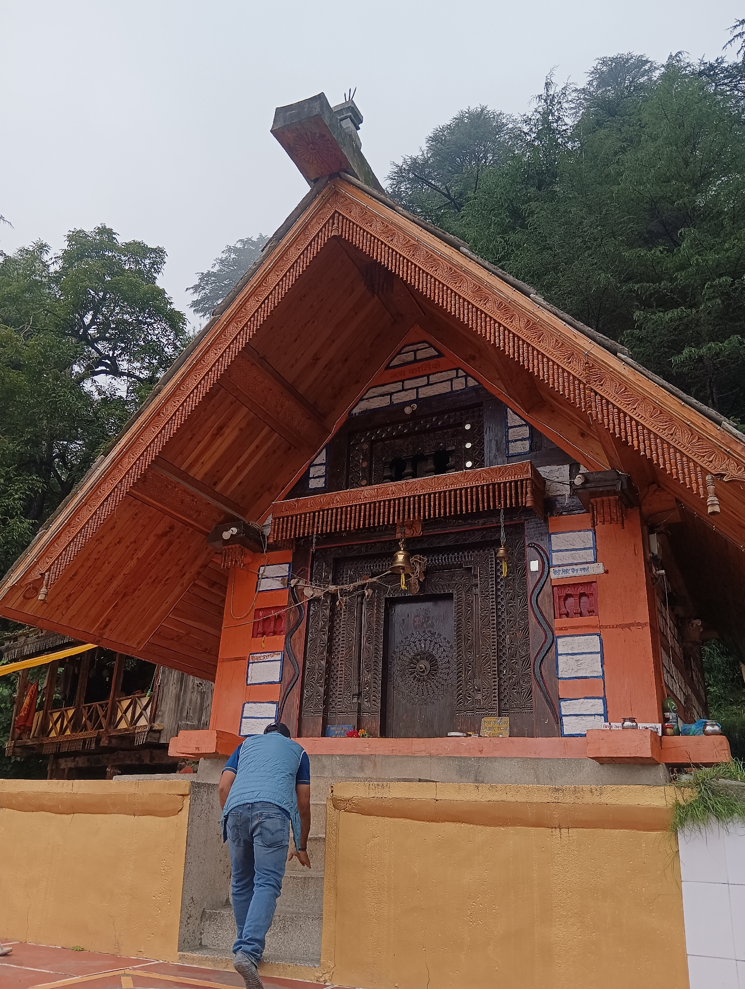 In Siumsa, India, a person walks up stone steps towards a wooden temple with an orange facade. The temple features intricate carvings on its large wooden doors and detailed wooden trim along its eaves. The roof is made of wood and has a pointed, gable design. The temple is situated on a hillside, with lush green trees forming a dense backdrop. The sky is overcast and the overall atmosphere is serene. The person is wearing a blue vest and jeans, their back is to the camera as they ascend the steps.
