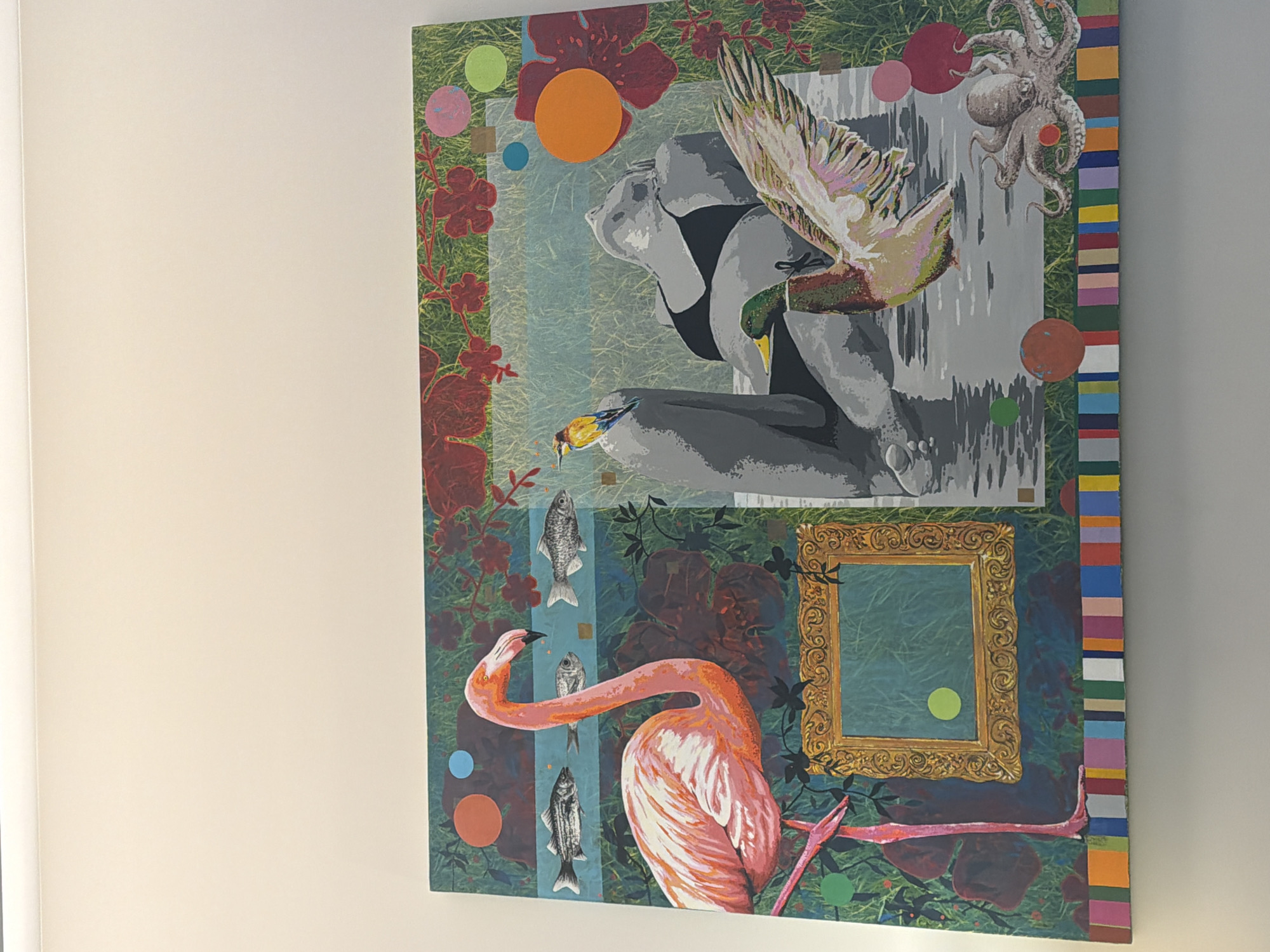 The image shows a vibrant and surreal painting displayed on a white wall, likely in a public or residential space. The painting is a large, horizontal canvas with a collage of various elements. On the left side, a large, bright pink flamingo stands prominently, its neck curved. Above the flamingo, three stylized fish swim in a horizontal line across a blue background. To the right of the flamingo, a decorative, ornate gold frame is depicted, with a green background inside it.
The right side of the painting features a grayscale depiction of a person reclining, wearing a black bikini top. A mallard duck with its wings spread is positioned in the foreground, appearing to emerge from water. Behind the reclining figure, a small bird with colorful plumage is perched on a gray, abstract rock formation. An octopus with multiple tentacles is shown at the bottom right of the painting.
The painting incorporates various circular shapes in different colors scattered throughout, and the overall background appears to be a lush green, suggesting a natural or garden-like setting, with red floral elements interspersed. The bottom edge of the painting is bordered by a colorful strip of vertical stripes. The lighting suggests an indoor environment with the painting illuminated from above. There are no specific landmarks or visual cues to indicate the city of Espinho, Portugal.