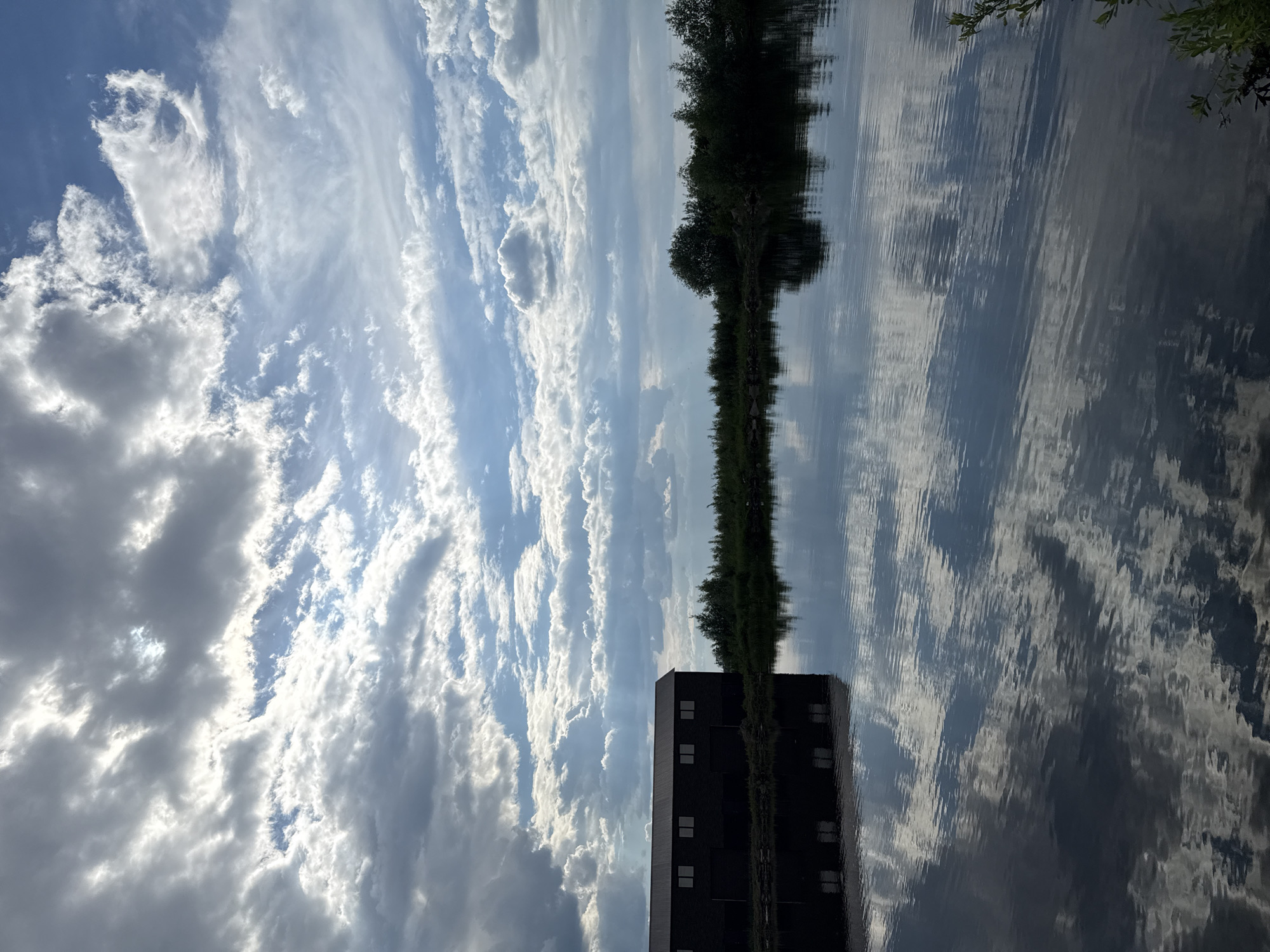 A wide shot captures a tranquil body of water reflecting a dramatic sky filled with dramatic clouds. On the left, a dark, rectangular building with multiple windows is silhouetted against the bright sky. The water's surface is disturbed by ripples, creating distorted reflections of the building and the clouds. The background features a dense line of green trees and foliage along the water's edge. The scene appears to be an outdoor setting, possibly a rural area or a park near a body of water. The lighting suggests it is late afternoon or early evening, with sunlight breaking through the clouds in some areas. The location is Chaikovsky, Russia, and this is likely a view of the Kama River or a similar large body of water within or near the city.