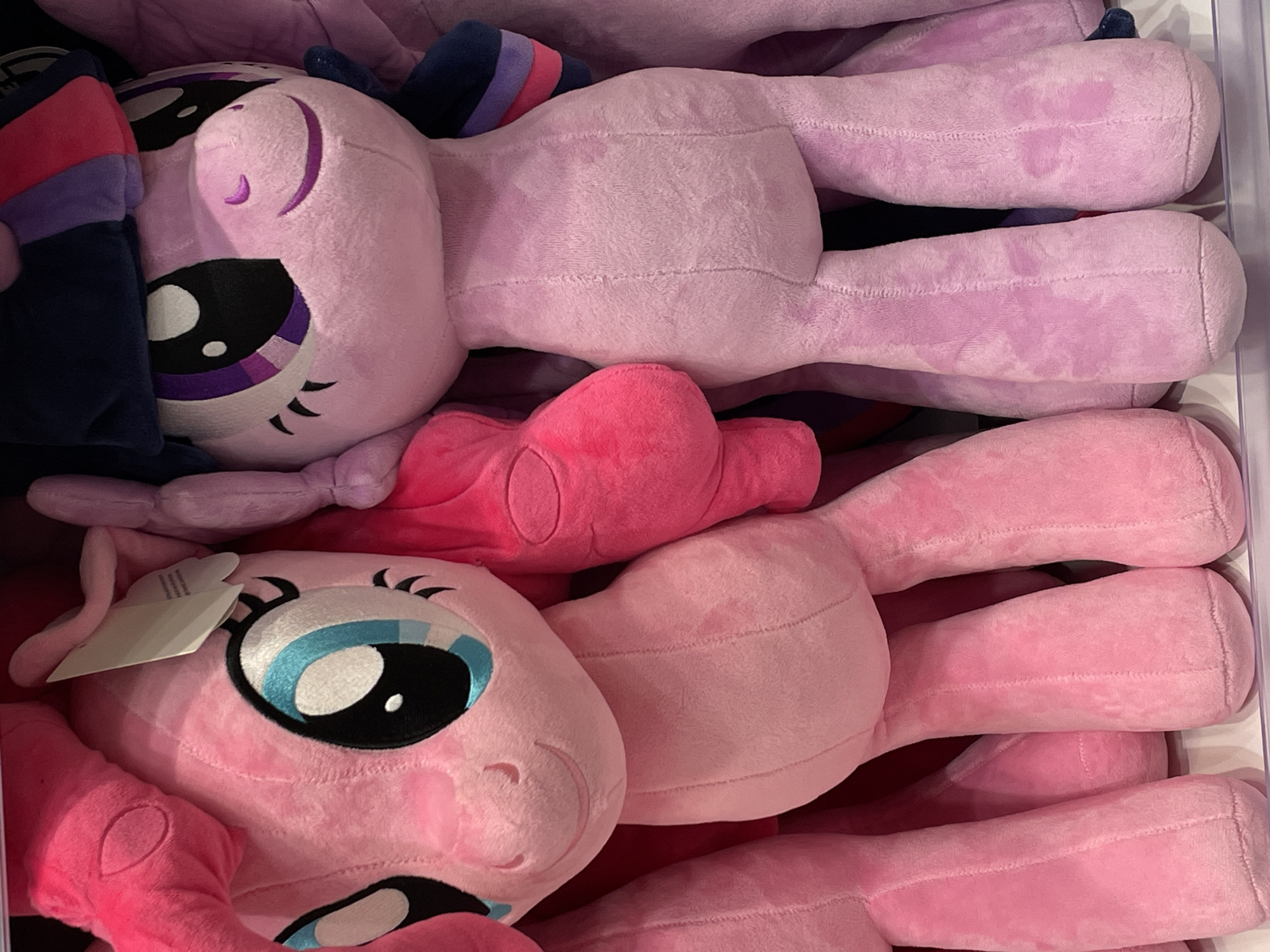 The image shows two large plush toys, identified as characters from "My Little Pony," positioned side-by-side on a shelf. The toy on the left is pink, representing the character Pinkie Pie, with large blue and black eyes, pink ears, and a pink mane. The toy on the right is lavender, representing the character Twilight Sparkle, with large purple and black eyes, a dark blue mane with purple and pink stripes, and dark blue ears. Both toys have their arms and legs extended, and are made of soft, fuzzy material. The background is blurred but suggests an indoor retail environment, possibly a toy store. The specific location within Davao City, Philippines cannot be determined from the image alone.