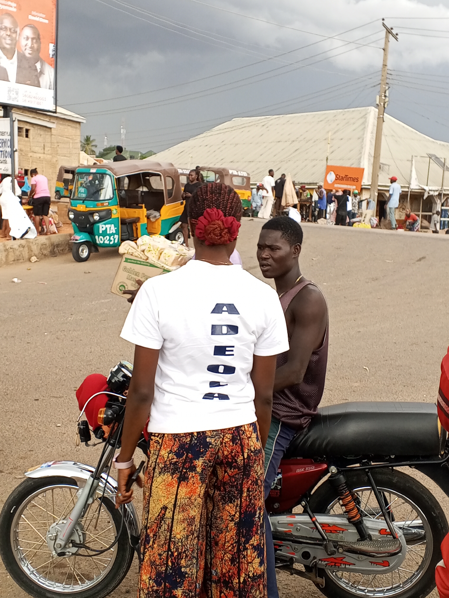 The image captures a scene in Gwong, Nigeria. A woman in a white t-shirt and patterned trousers stands next to a man sitting on a parked motorcycle. She has red braids styled on the back of her head and carries a box of baked goods. The man, wearing a sleeveless shirt, is in conversation with the woman.
Behind them, there are several yellow and green auto-rickshaws (tuk-tuks). A crowd of people is gathered in the background. A billboard, a street, and a StarTimes sign are also visible. The sky is overcast with heavy clouds.