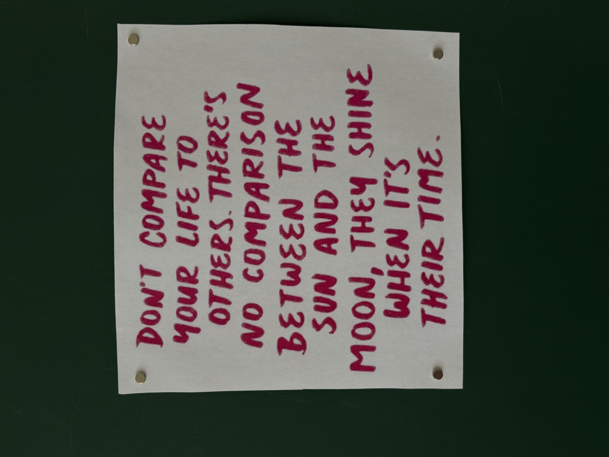A piece of off-white paper is affixed to a dark green surface using four silver push pins. The paper contains red text arranged in vertical lines reading, "DON'T COMPARE YOUR LIFE TO OTHERS. THERE'S NO COMPARISON BETWEEN THE SUN AND THE MOON, THEY SHINE WHEN IT'S THEIR TIME." The paper is slightly creased or unevenly cut. The dark green surface appears flat and matte. The scene is indoors with even lighting.