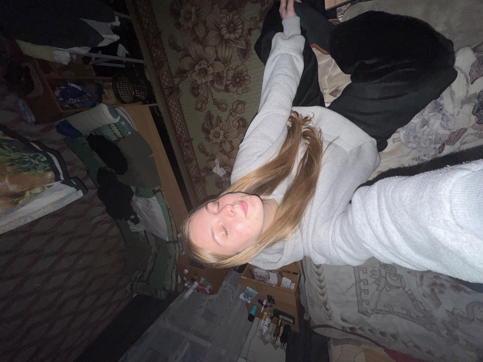 A young fair-skinned individual with blonde hair, wearing a light grey hoodie and dark pants, is lying on patterned bedding and a floral rug. The individual's eyes are closed, and they are facing towards the upper right corner of the frame. The setting appears to be a room with various items scattered around, including a shelving unit with clothes, small bottles on a wooden surface, and miscellaneous items on the floor. The image's perspective suggests it was taken from a high angle, possibly a selfie. The location appears to be a residential interior, presumably in Perm, Russia.