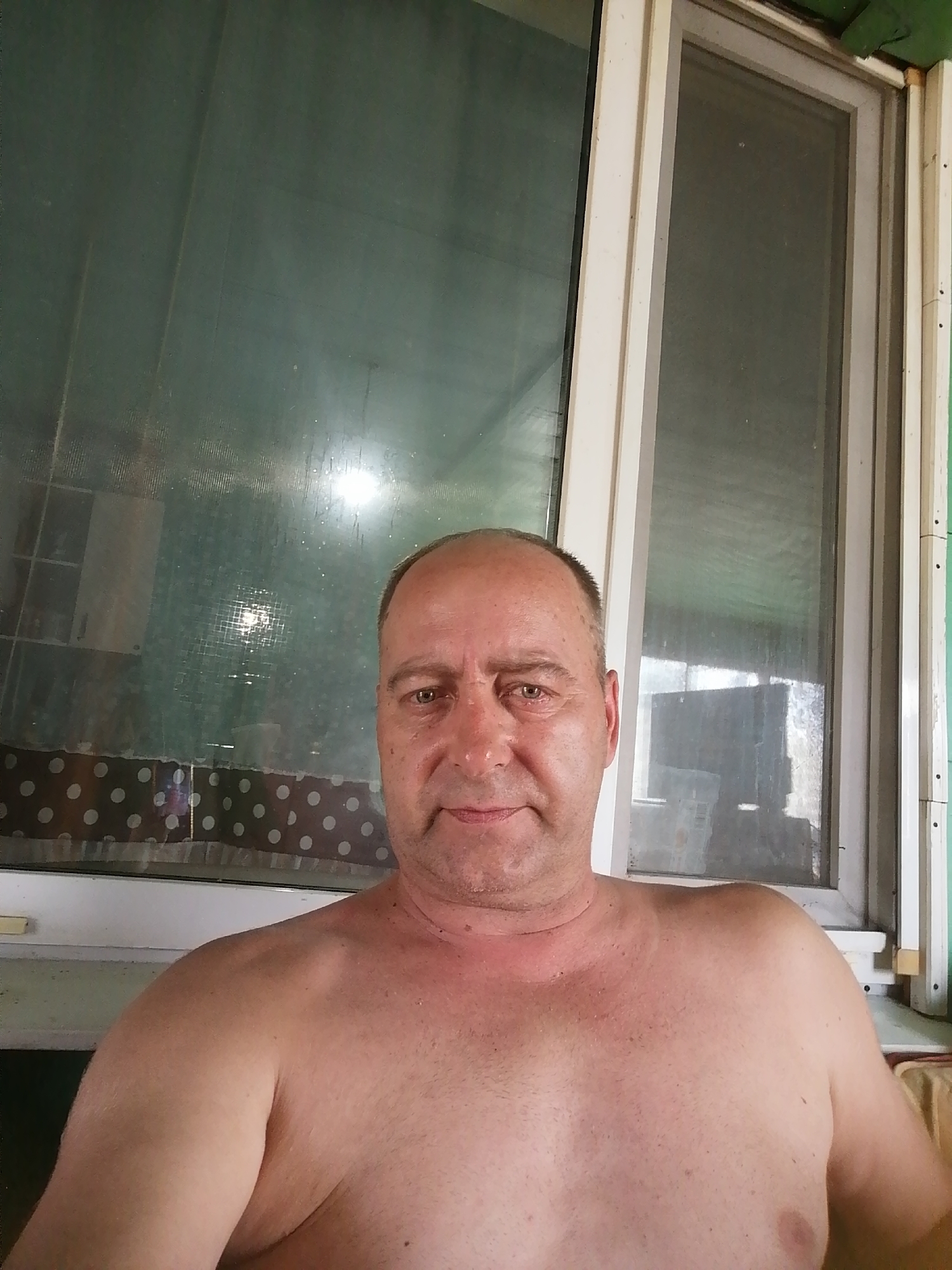 A middle-aged Caucasian man with a bald head and fair skin is shown from the chest up, facing the camera. He is shirtless and his skin appears slightly flushed. His expression is neutral. He is seated in front of a window with two panes. The left pane is dark and reflects light, showing some objects inside. The right pane is lighter and shows a reflection of what appears to be a room. A white window frame separates the two panes and extends to the sides. A patterned curtain with white polka dots on a dark background is visible below the window. The overall scene appears to be an indoor setting, possibly a porch or room with a view of the outdoors. The photograph was taken in Novopokrovka, Russia.