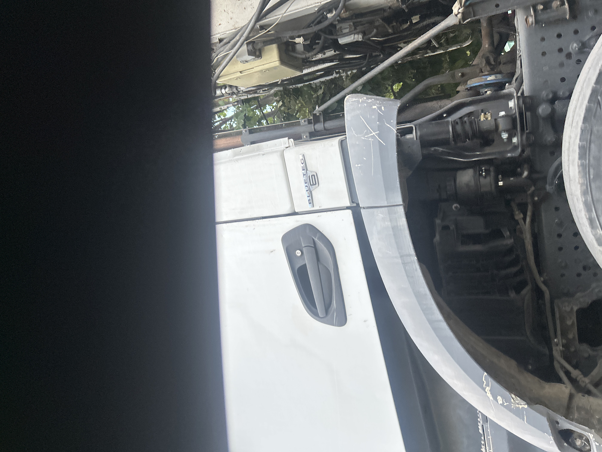 The image shows a close-up of a white truck's side door and wheel arch, with a black, blurry foreground element obscuring the top portion of the frame. The truck features a dark gray door handle with a lock cylinder. Below the door, a "BLUETEC 6" emblem is visible. The wheel arch is also dark gray and shows some scuff marks. Visible mechanical components and wiring are present behind the wheel arch. Green foliage is visible in the background, suggesting an outdoor setting, potentially in Varna, Bulgaria, given the provided context. No people are depicted in the image.