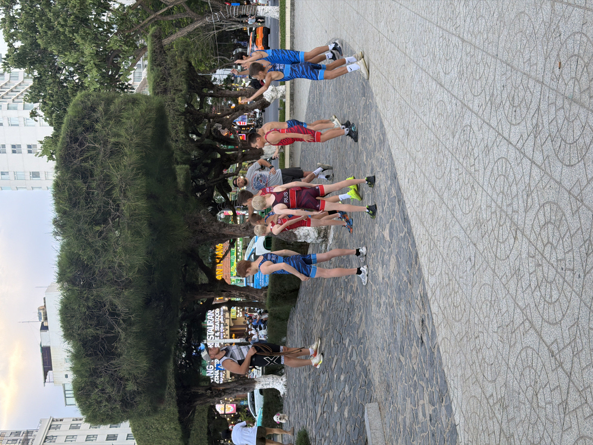 The image depicts an outdoor scene in Nha Trang Ward, Vietnam, likely a public park or plaza, characterized by paved ground with a circular patterned design. In the foreground, a group of young boys, some wearing wrestling singlets with "RUS" printed on them, are standing and interacting. One boy in a blue and black singlet with "RUS" on it is facing away from the camera, with his left arm raised. Another boy to his right is also in a blue singlet and has his right arm raised. To the left, a man in a grey and black striped tank top and a baseball cap stands holding a rope, facing a group of boys. Another man in a grey t-shirt is visible in the background, seemingly observing. In the middle ground, there are trees with distinctive rounded canopies and several people walking or standing. Behind the trees, commercial buildings, including a building with "RESTAURANT" visible on a sign, are present, suggesting an urban setting. Vehicles, including cars and motorcycles, are also visible on a street in the background. The overall atmosphere appears to be casual, possibly related to a sports training or gathering. A small white dog with a pink accessory on its back is also present on the paved area. The sky is overcast, indicating a cloudy day.