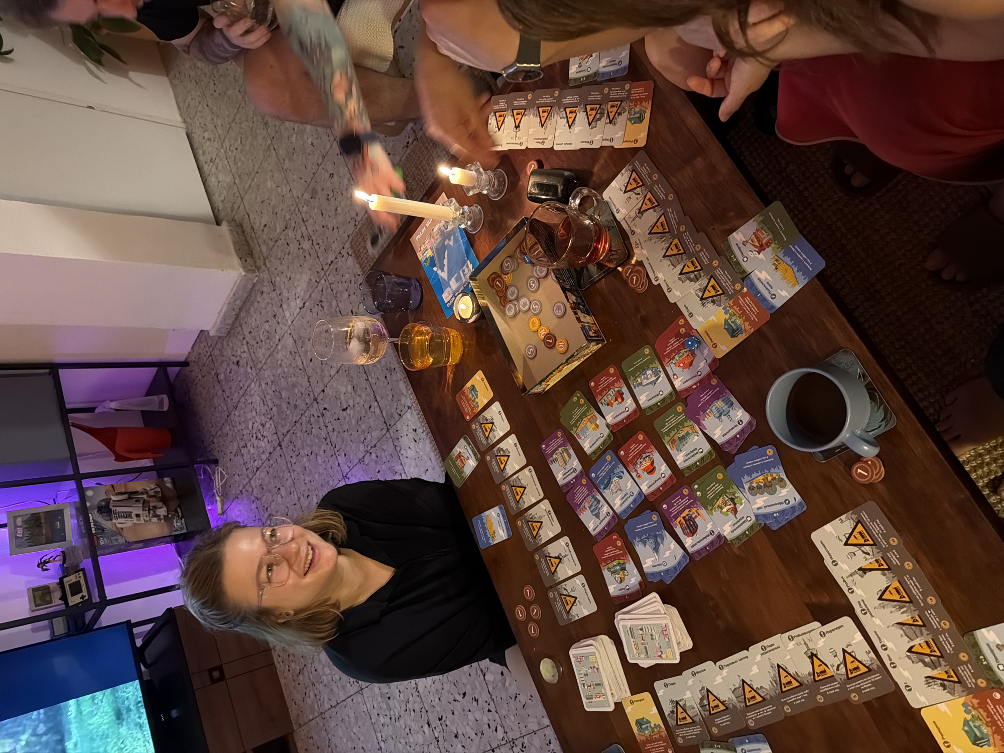 The scene depicts a group gathered around a wooden table indoors, engaged in a board game. The focus of the gathering is a woman with blonde hair and glasses, smiling at the camera from the left side of the frame. The table is covered with numerous game cards, coins, a box, and a mug, along with beverages in glasses. Candles provide additional lighting on the table, casting light on a male arm with a tattoo reaching across. Various individuals are situated around the table, with only parts of their bodies visible. The location is a home, with a television and shelving unit in the background. The location could be Ramat Gan, Israel.