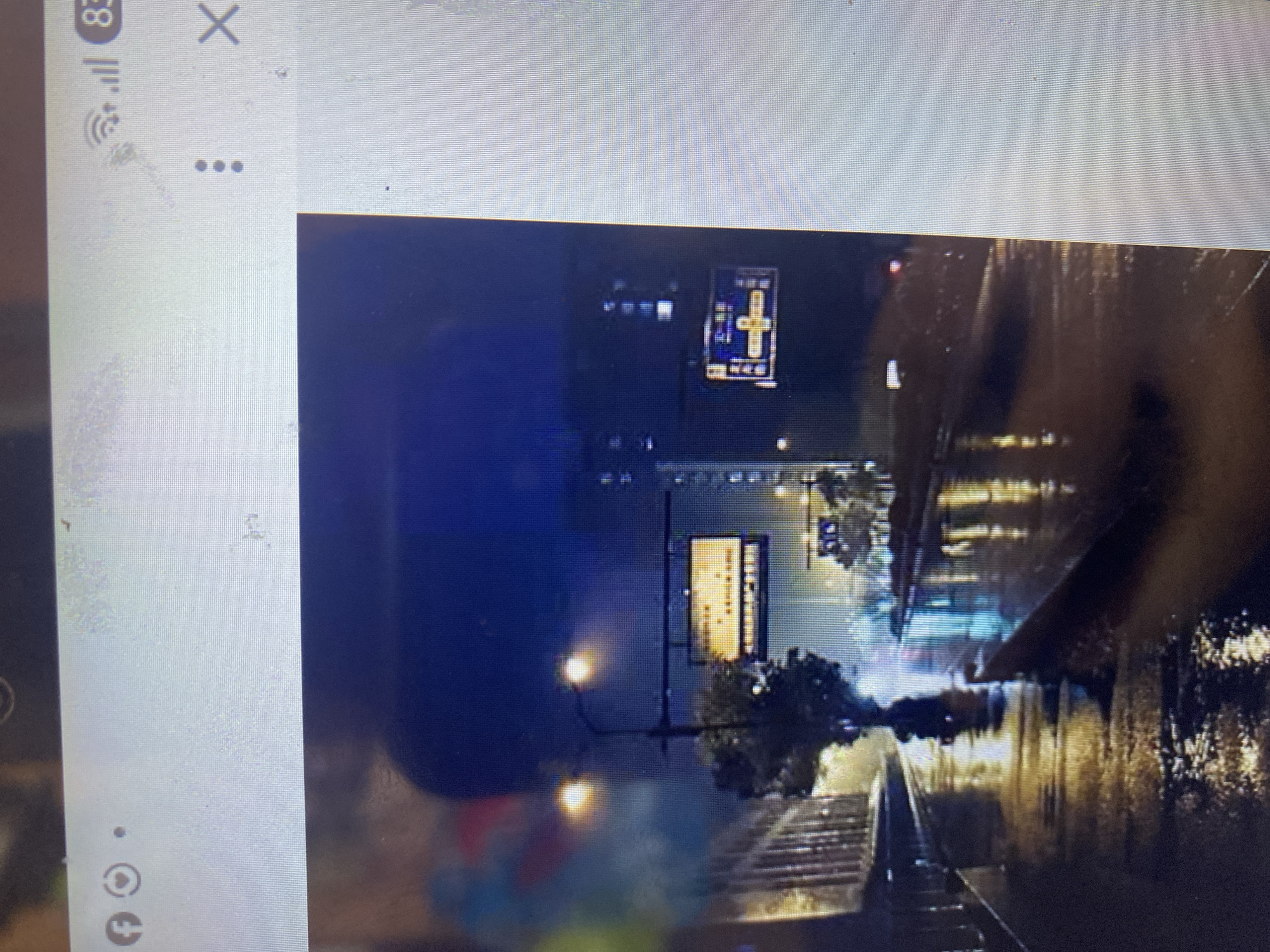 The image shows a nighttime scene in a city, likely Ta', Indonesia. The street is wet and reflects the lights from buildings and street lamps, suggesting it has been raining or is currently raining. There are traffic lights visible, as well as illuminated signs. A tree is situated on the left side of the road, and a building with glass windows is also visible. The overall atmosphere is dark and moody, with the reflections on the wet surface adding to the visual texture. The image appears to be captured from inside a vehicle or from a slightly elevated position, looking down a street.