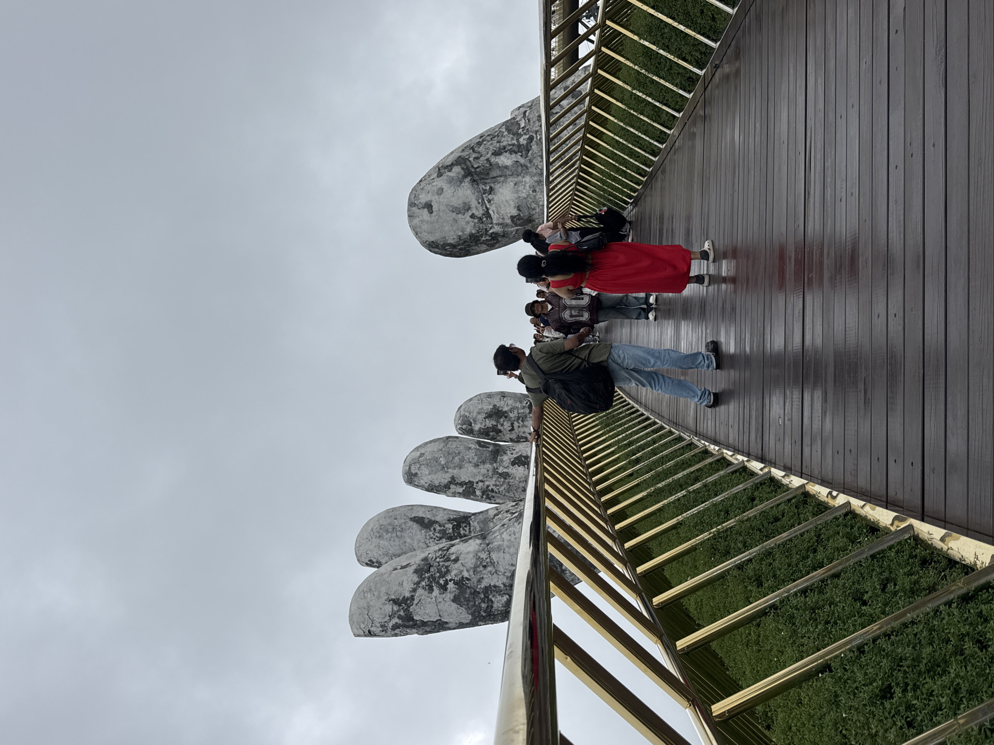 A low-angle shot depicts a section of the Golden Bridge, also known as the Cầu Vàng, in Đà Nẵng, Vietnam. The bridge features a wooden walkway, bordered by a gold-colored metal railing. Beneath the railing is a patch of dense, green vegetation. The bridge is supported by two enormous stone hands, which appear aged and weathered. Several people are visible on the bridge. Some individuals are taking photographs with their phones or cameras. One person is wearing a bright red dress. The sky is overcast and gray, suggesting cloudy weather conditions. The surface of the wooden bridge appears to be wet, possibly from rain.