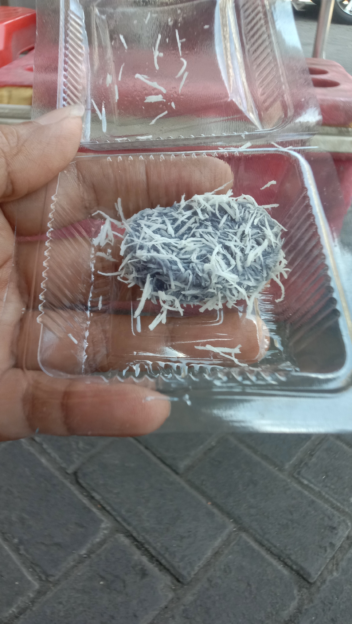 A close-up, high-angle shot shows a person's hand holding a clear plastic container with a purple sweet treat covered in shredded white coconut. The sweet is oval-shaped and appears to be a rice cake or a similar confection. The container is semi-transparent, revealing the texture of the sweet beneath. The background is blurred, but suggests an outdoor setting with paving stones visible at the bottom of the frame. The lighting is bright, casting subtle shadows.