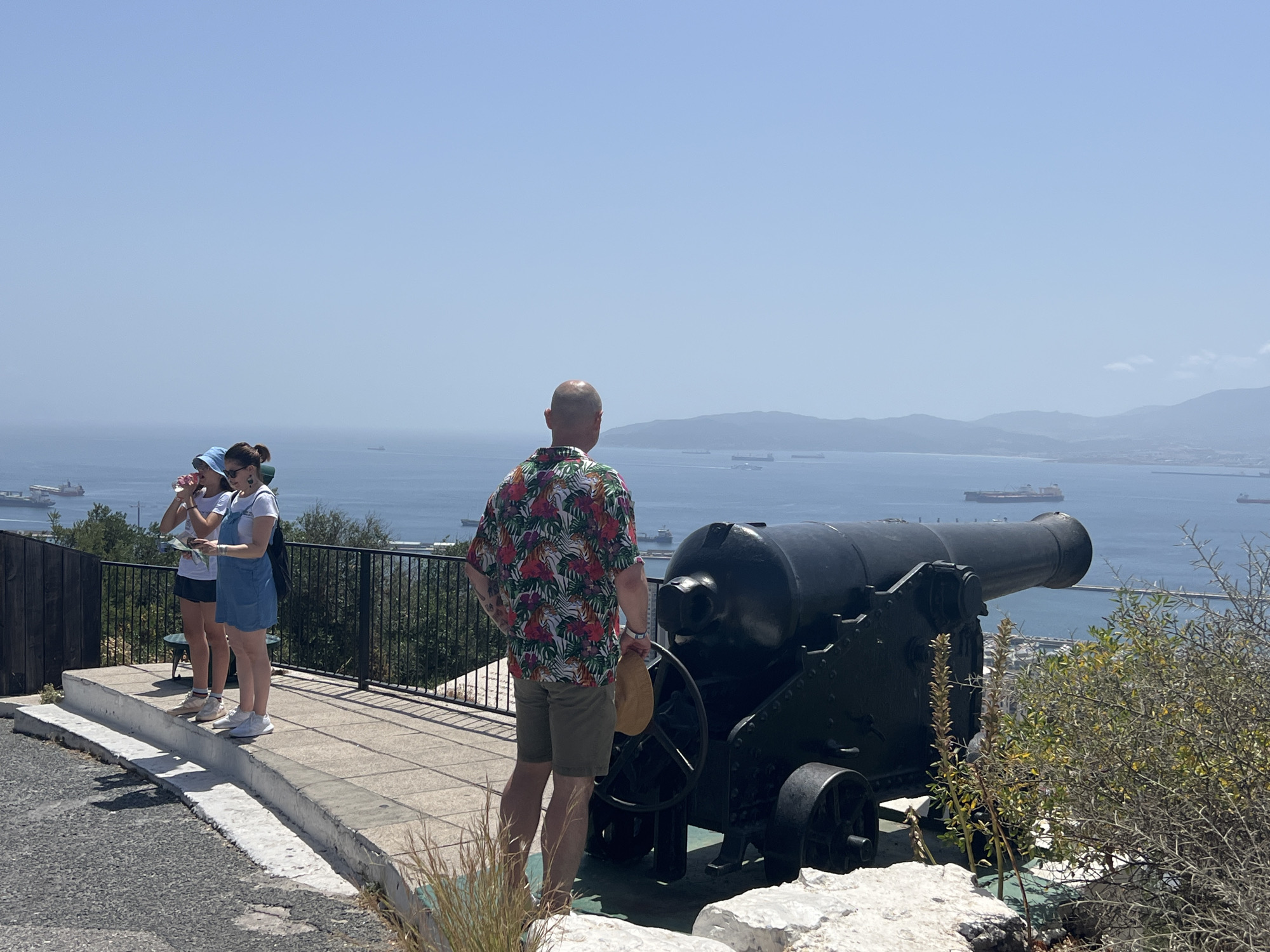 The image depicts a sunny day on a raised observation point overlooking the sea in Gibraltar. A large, black cannon is prominently displayed in the foreground, positioned to face the expansive blue water where several cargo ships are visible. To the left, three individuals are standing on a tiled walkway behind a black metal railing. Two of them, appearing to be young women, are facing the sea, with one holding a pink object to her mouth and the other looking at a piece of paper. A man wearing a brightly colored, floral-patterned short-sleeved shirt and khaki shorts stands with his back to the camera, holding a tan hat in his right hand and looking out at the same view. The environment is characterized by a clear sky, the shimmering sea, and hints of distant landmasses. There is a weathered concrete wall in the lower left of the image, and some dry vegetation grows in the lower right corner. The overall scene suggests a historical or scenic tourist attraction in Gibraltar.